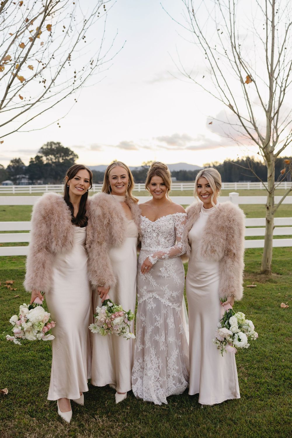 Wedding party in blush pink and white gowns stand together holding bouquets against a sunset countryside backdrop.