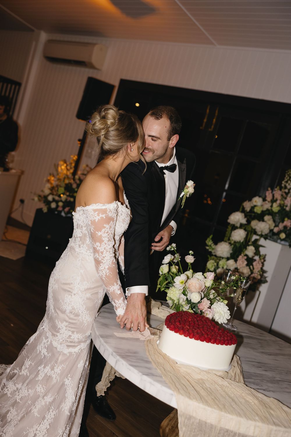 A romantic couple in wedding attire leaning together near a white and red cake at their reception.
