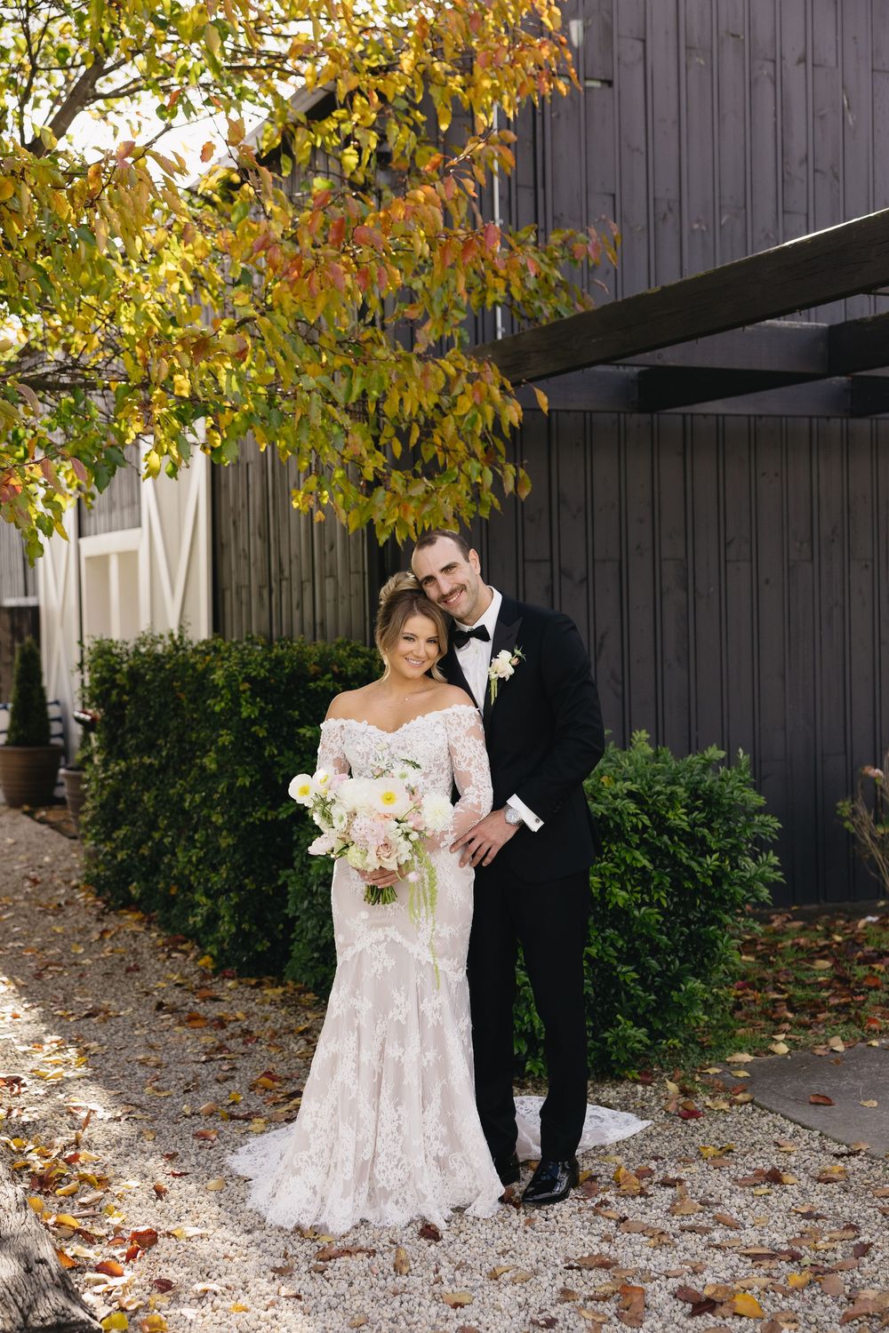 A wedding couple poses against a dark barn wall in autumn with golden leaves and fallen foliage.