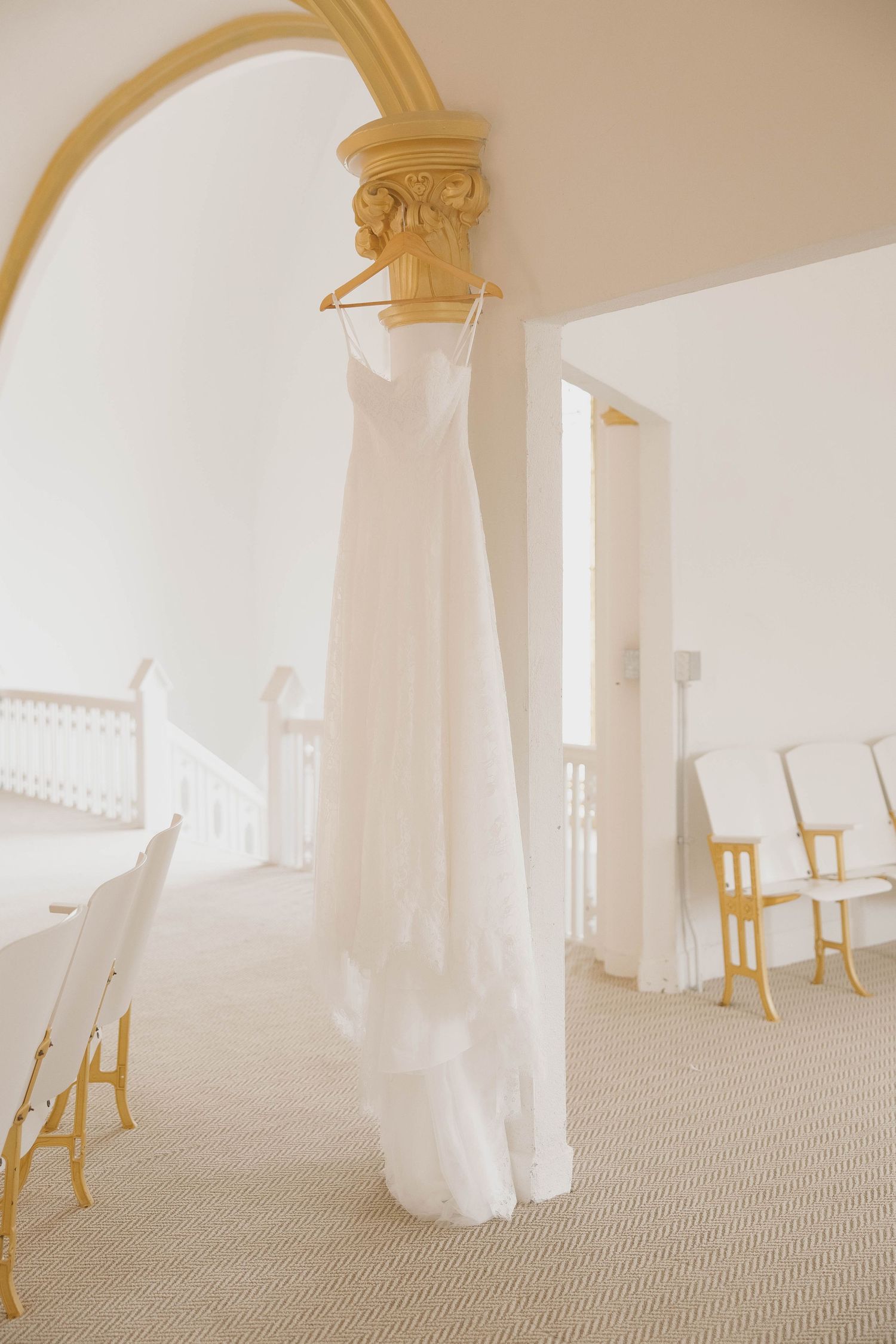 Elegant white wedding dress hanging from ornate architectural column in bright sunlit room with beige carpet.