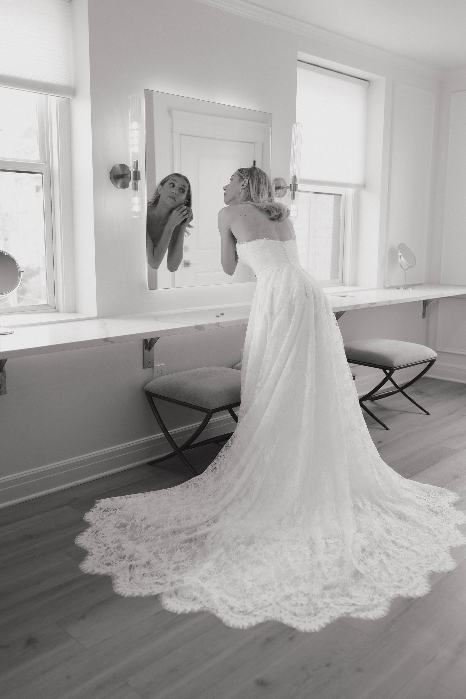 Black and white photo of a bride in an elegant lace wedding gown with long train looking in mirror at dressing room.