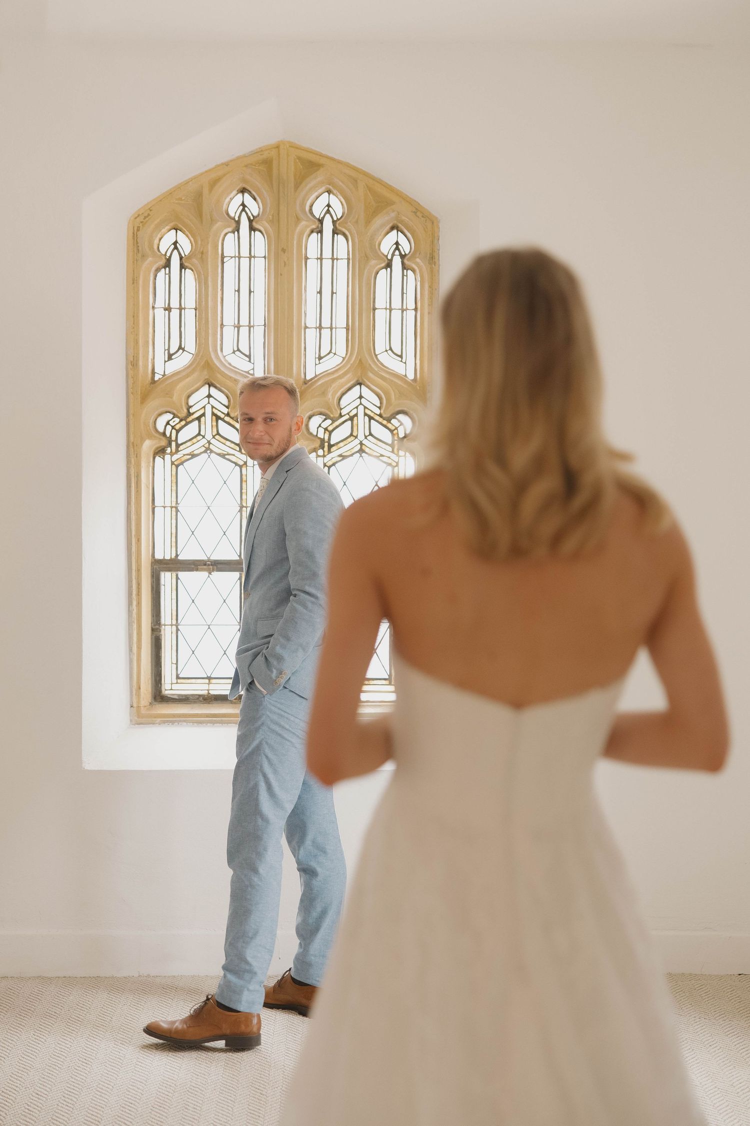 A romantic first look moment between couple in light-filled chapel with gothic stained glass windows and white walls.