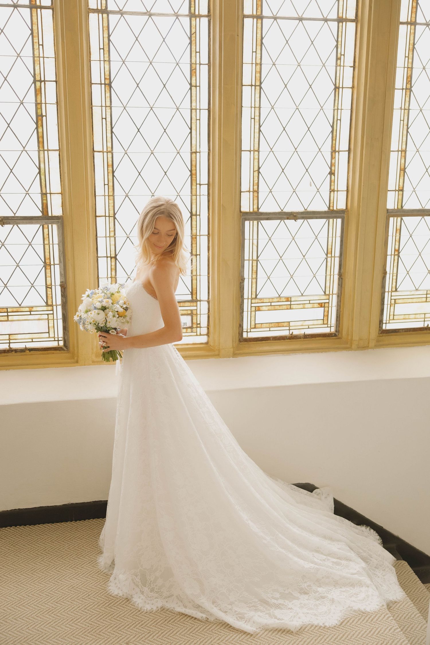 A bride in an elegant white strapless gown with a flowing train poses by stained glass windows holding a white bouquet.