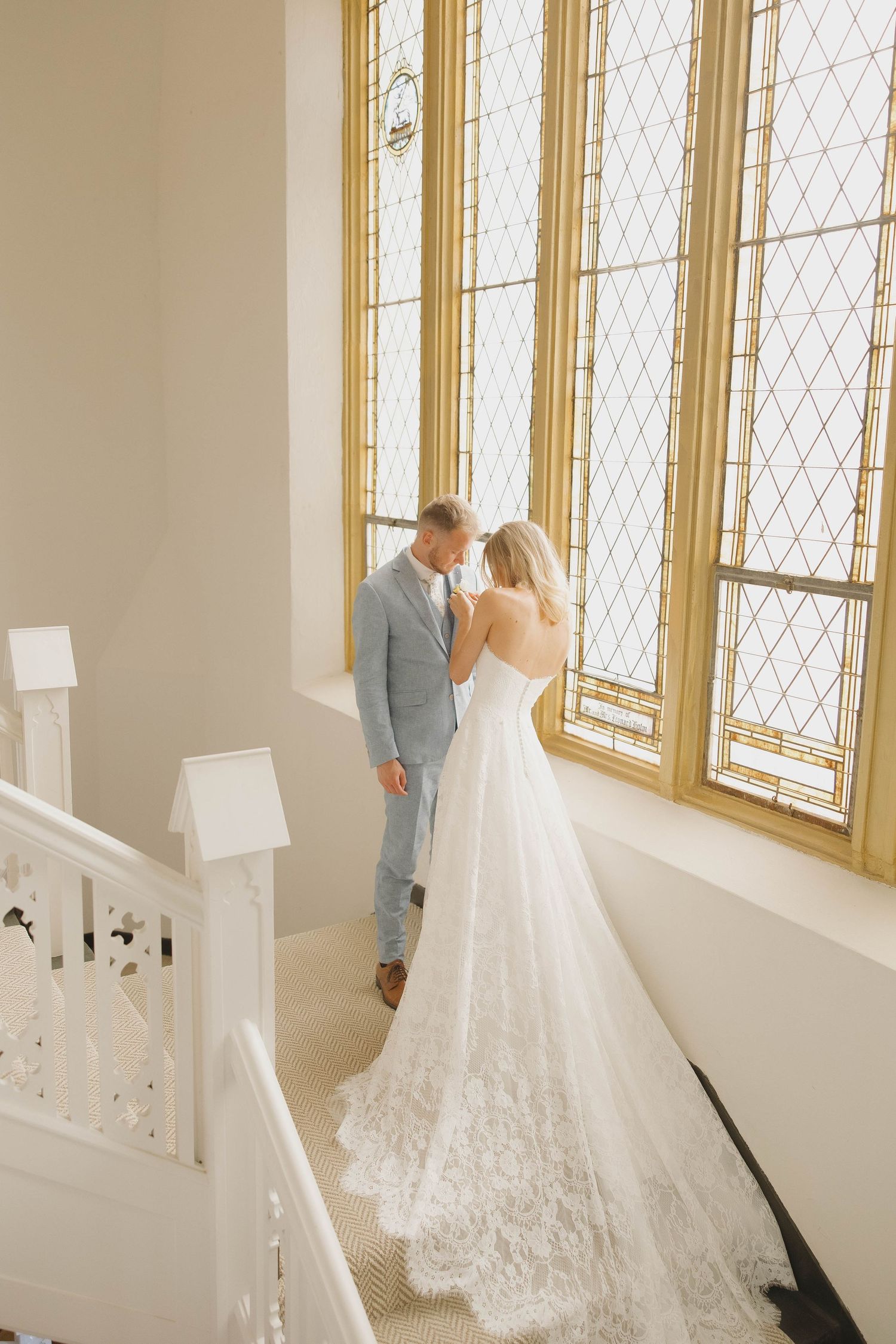 A couple in wedding attire stands by large ornate windows with diamond-patterned glass in an elegant white interior stairway.