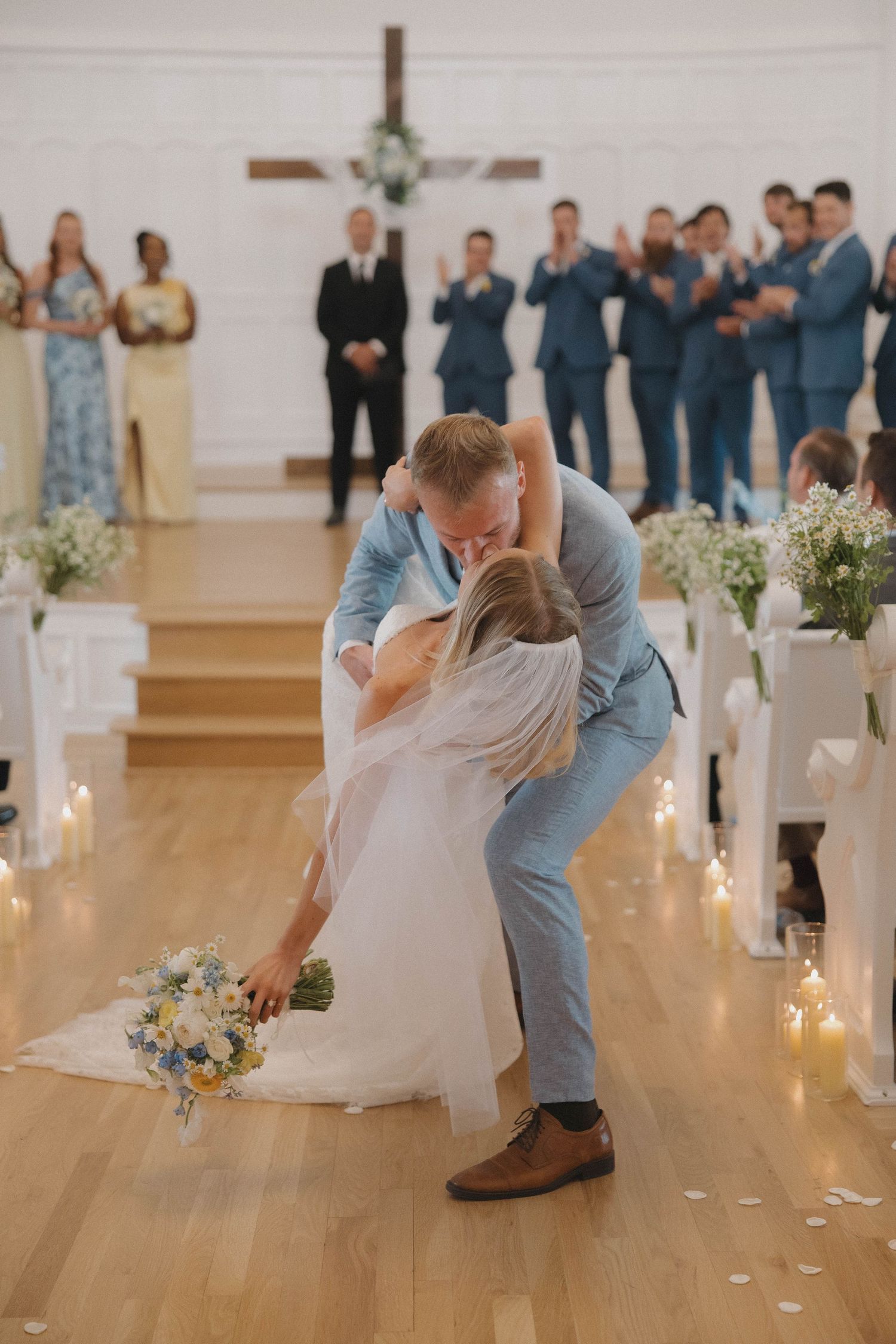 Newlyweds share romantic dip kiss in church aisle with wedding party and floral decorations in background.