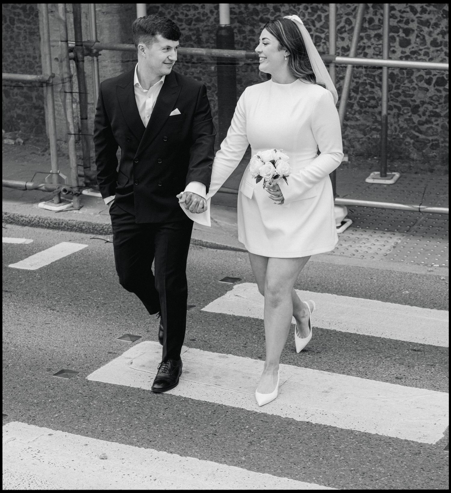 A couple walking together on a zebra crossing, with the bride in a short white dress and groom in a dark suit.