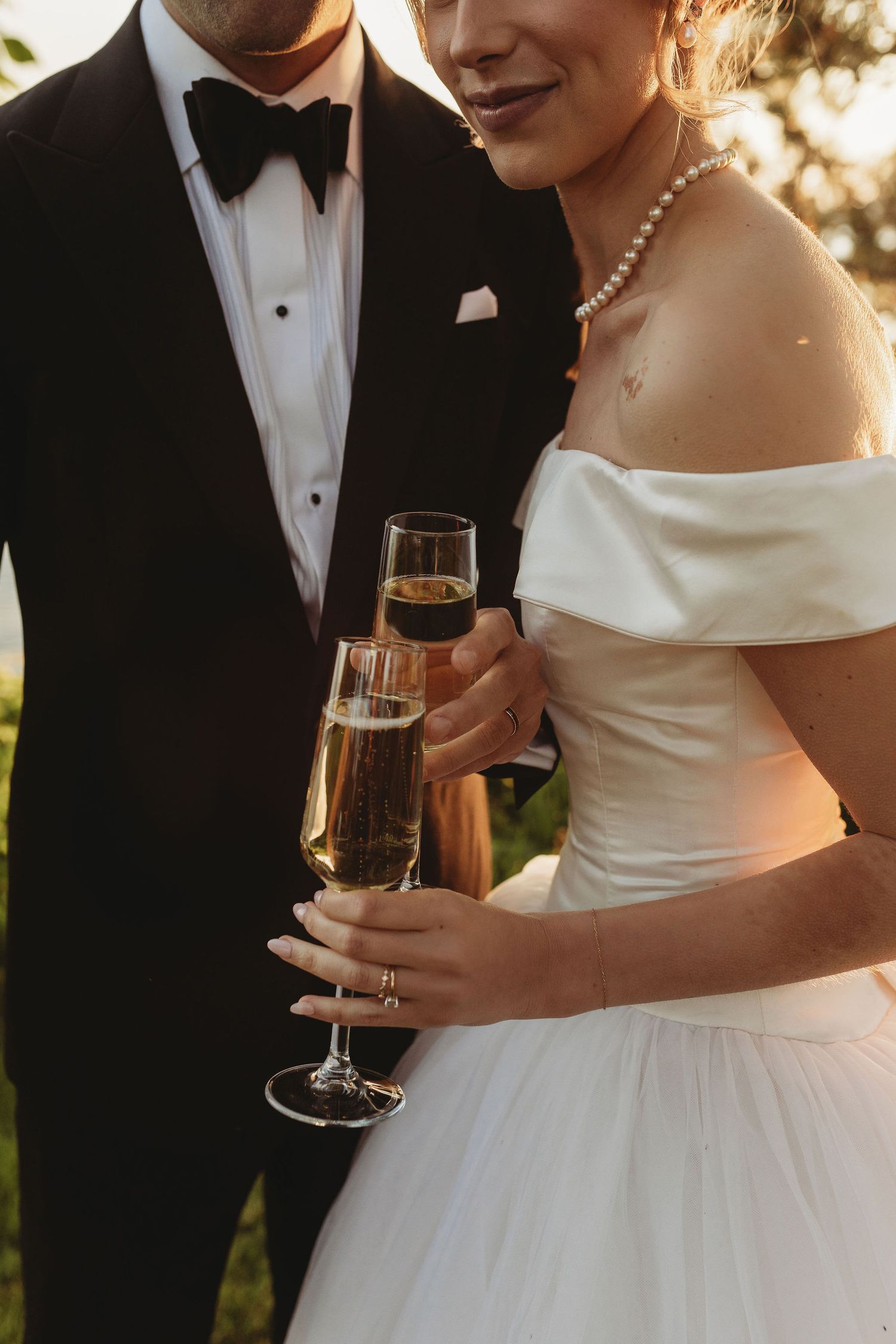 Close-up details of a bride in white gown and groom in tuxedo toasting with champagne flutes at sunset.