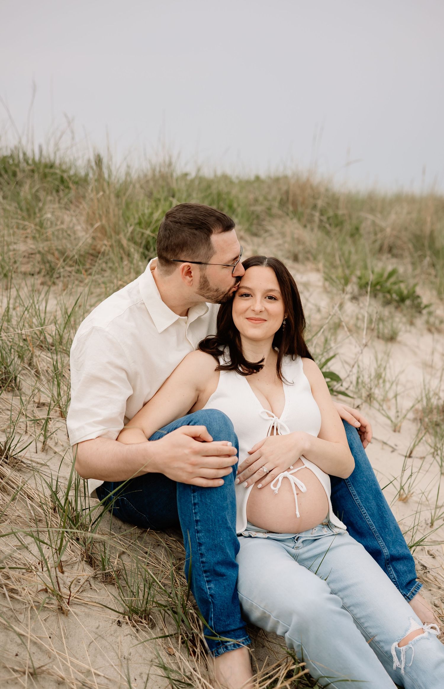 Couple sits together intimately at sandy beach dunes wearing casual white and denim outfits during maternity photoshoot.