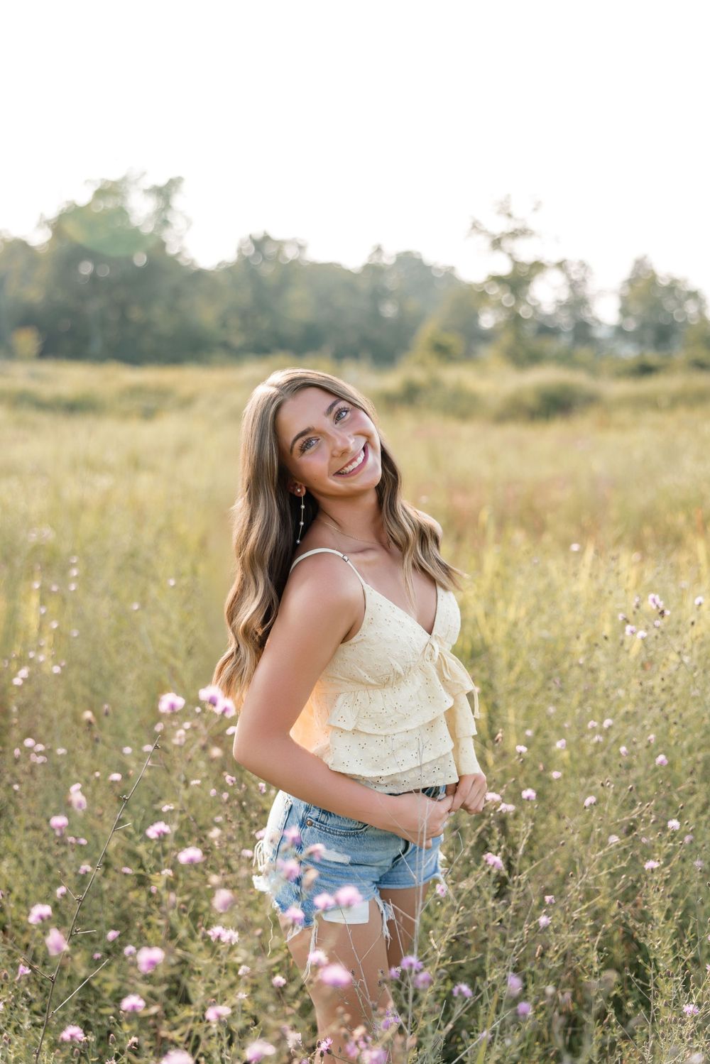 A person in a white tank top and denim shorts poses joyfully in a wildflower meadow at golden hour.