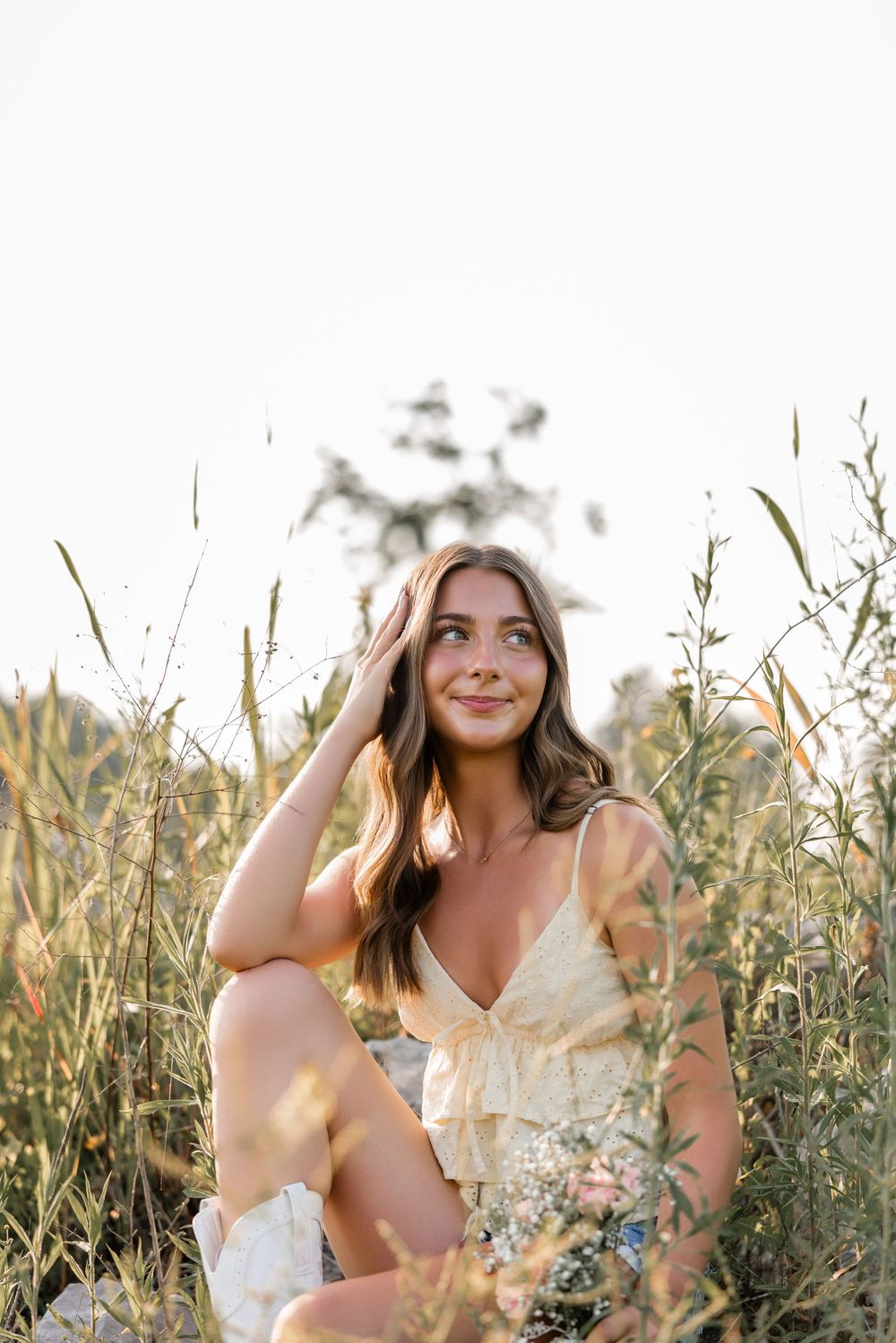 Woman in light dress sits dreamily among tall grass and wildflowers in golden sunlight.