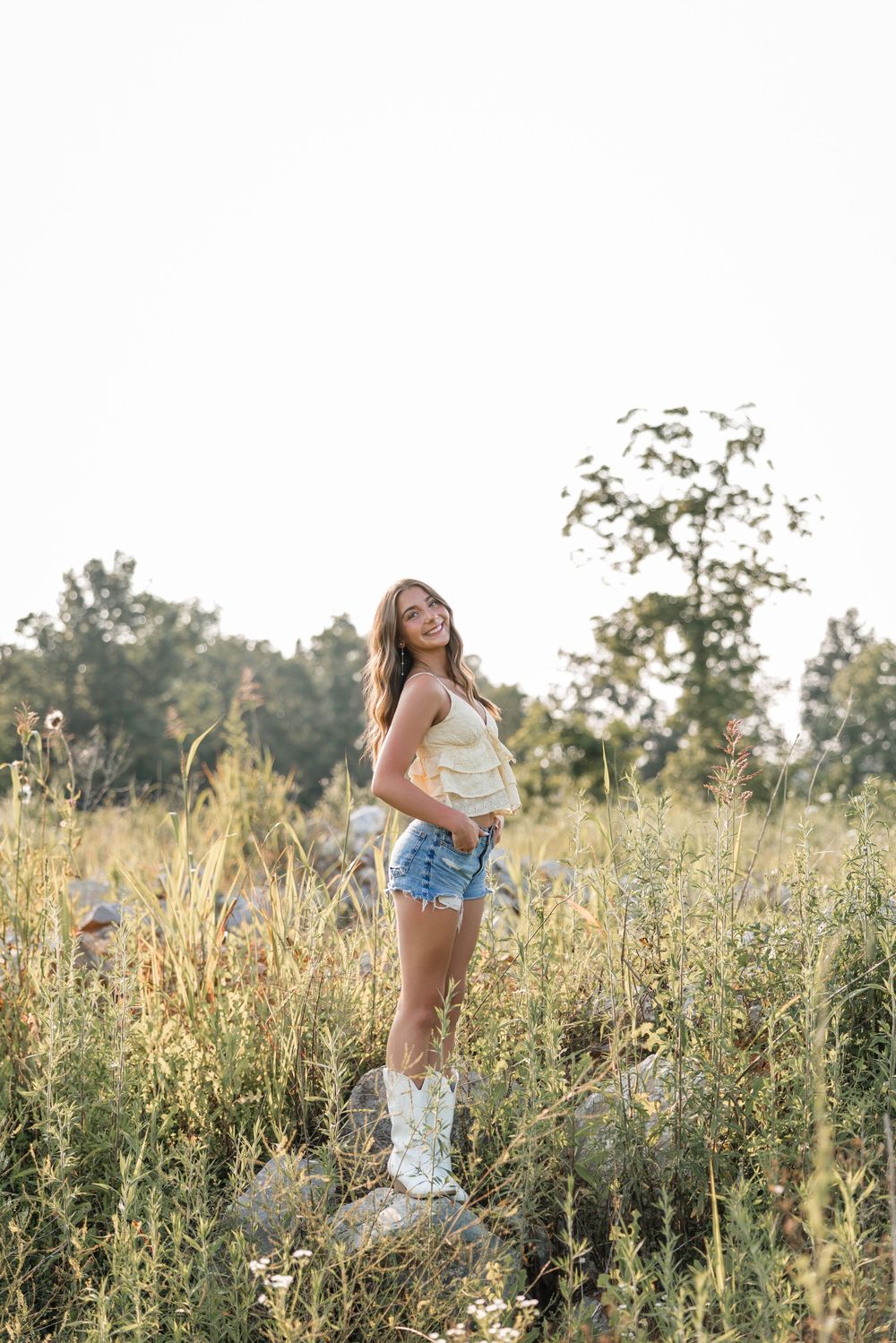Person wearing white cowboy boots and denim shorts poses in a sun-lit wildflower field during golden hour.