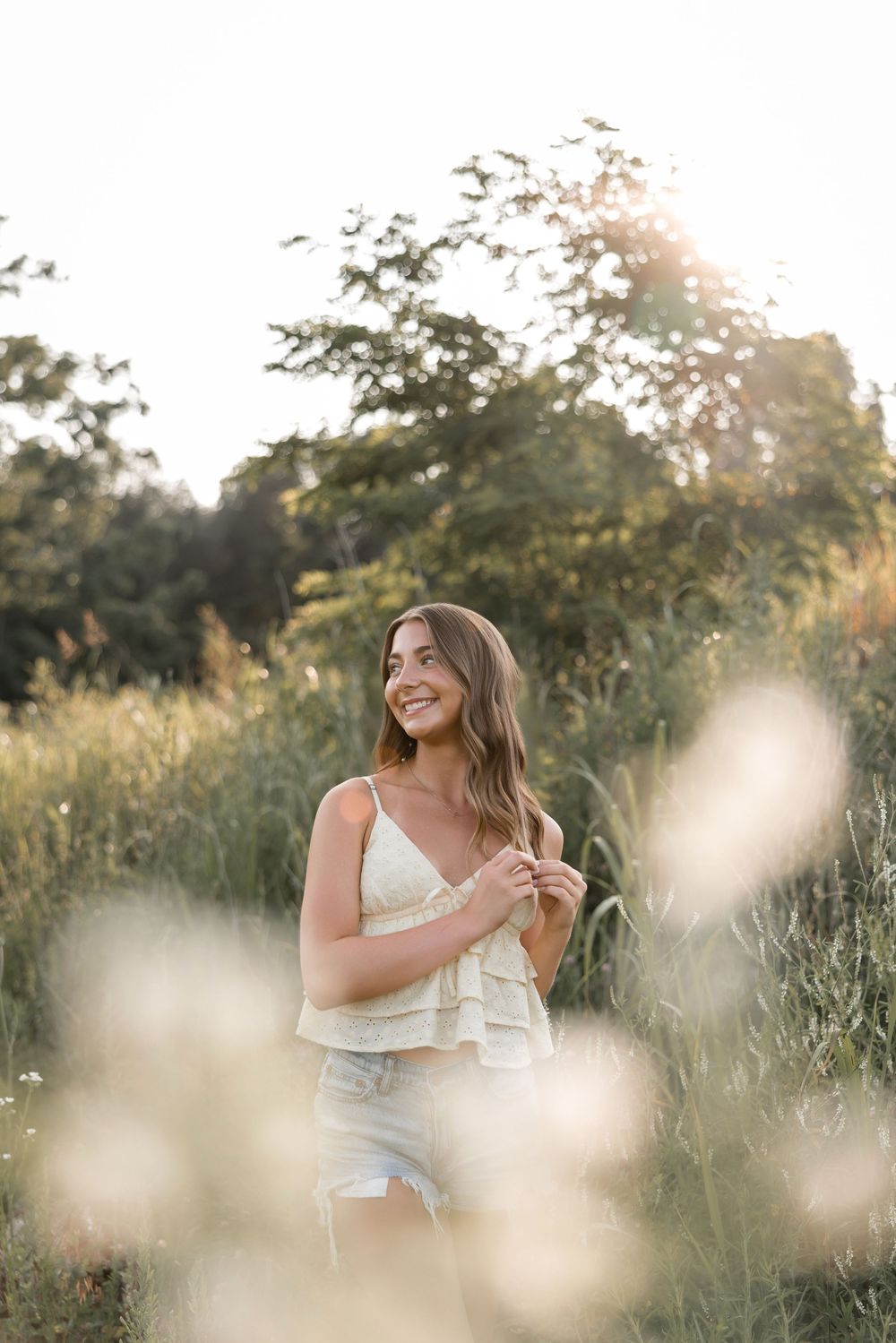 A person in a white summer top stands joyfully in a sunlit meadow during golden hour with soft bokeh in foreground.
