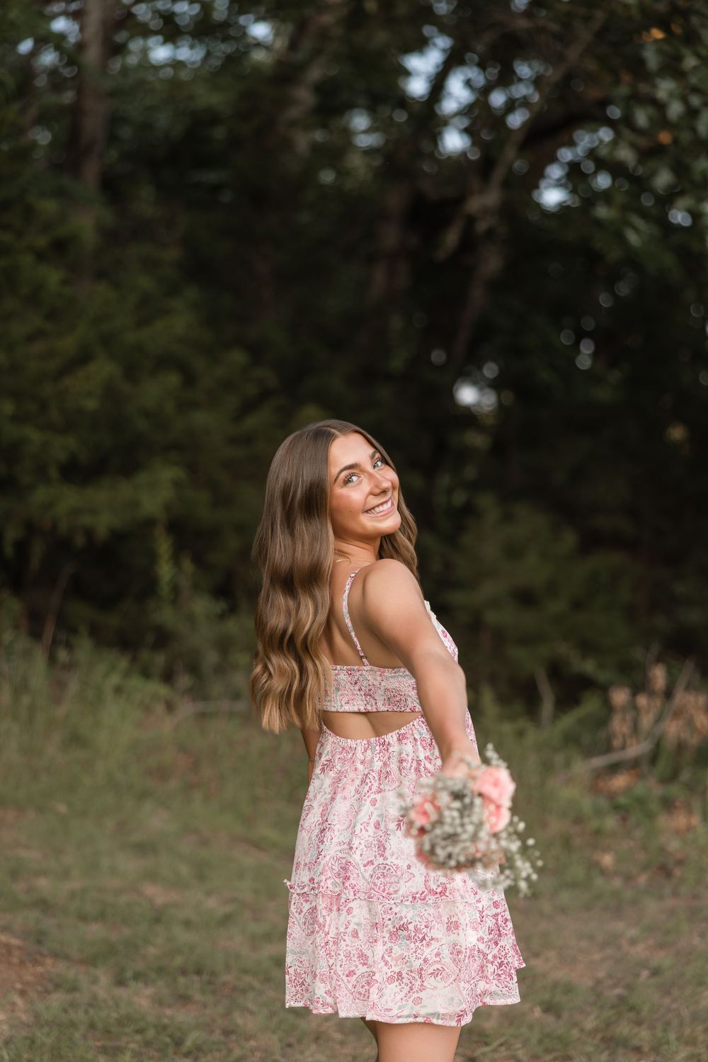 Person in pink floral dress holds wildflower bouquet while standing in outdoor wooded setting during summer.