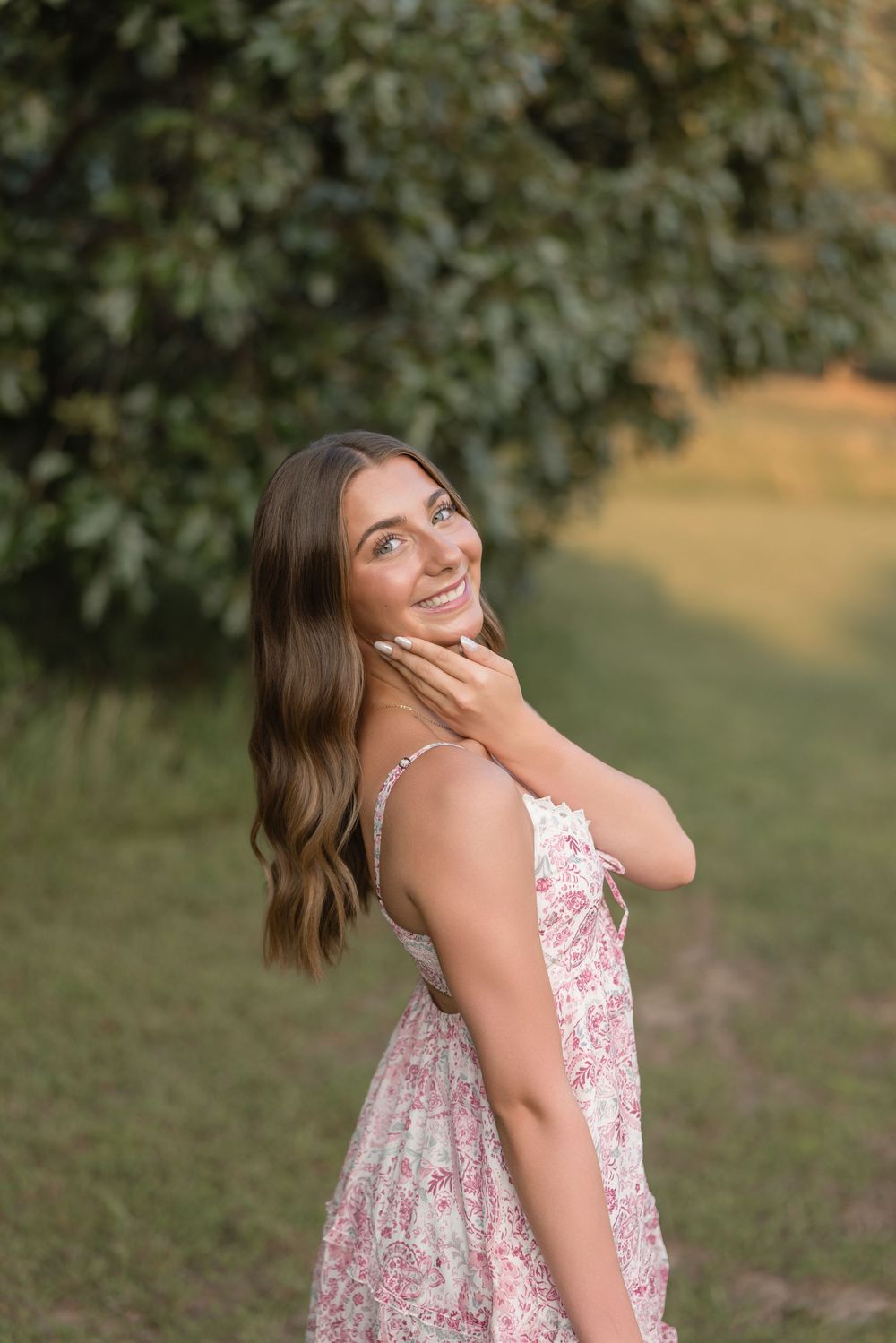 A person in a floral sundress laughs joyfully while standing outdoors near trees during a warm summer day.