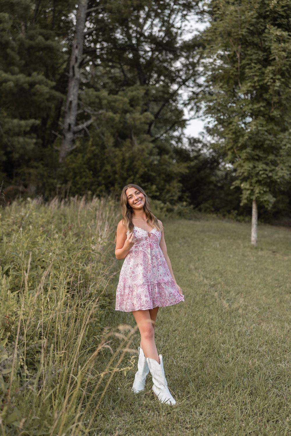 A person in a pink floral dress poses outdoors in a field with trees in the background on a sunny day.