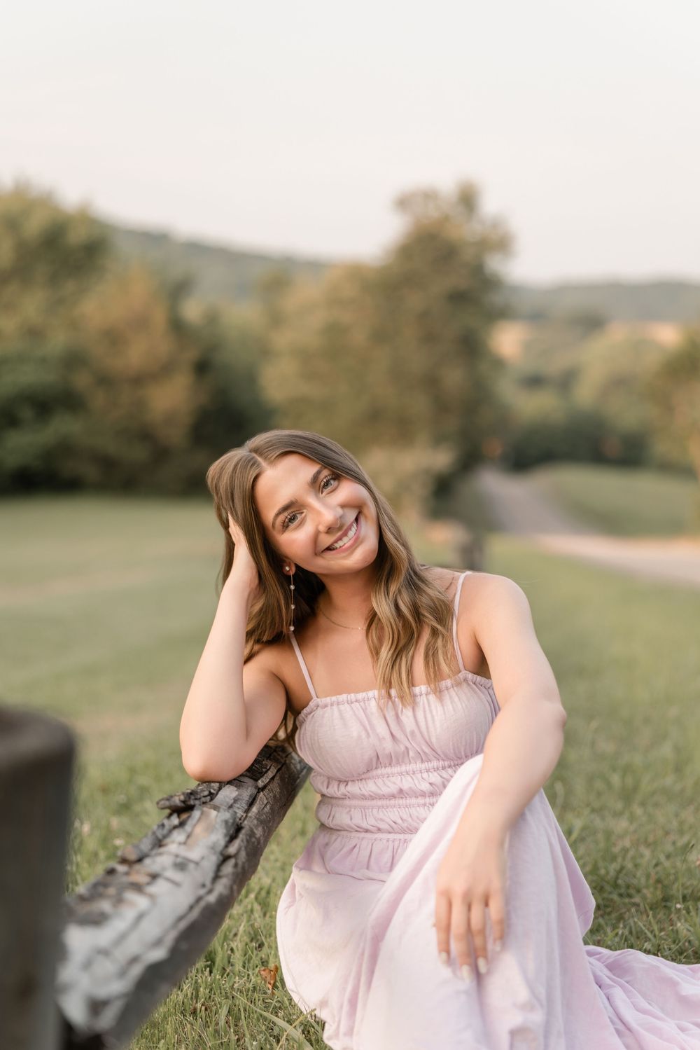 A woman in a light pink dress sits on grass with rolling hills and trees in the background during golden hour.