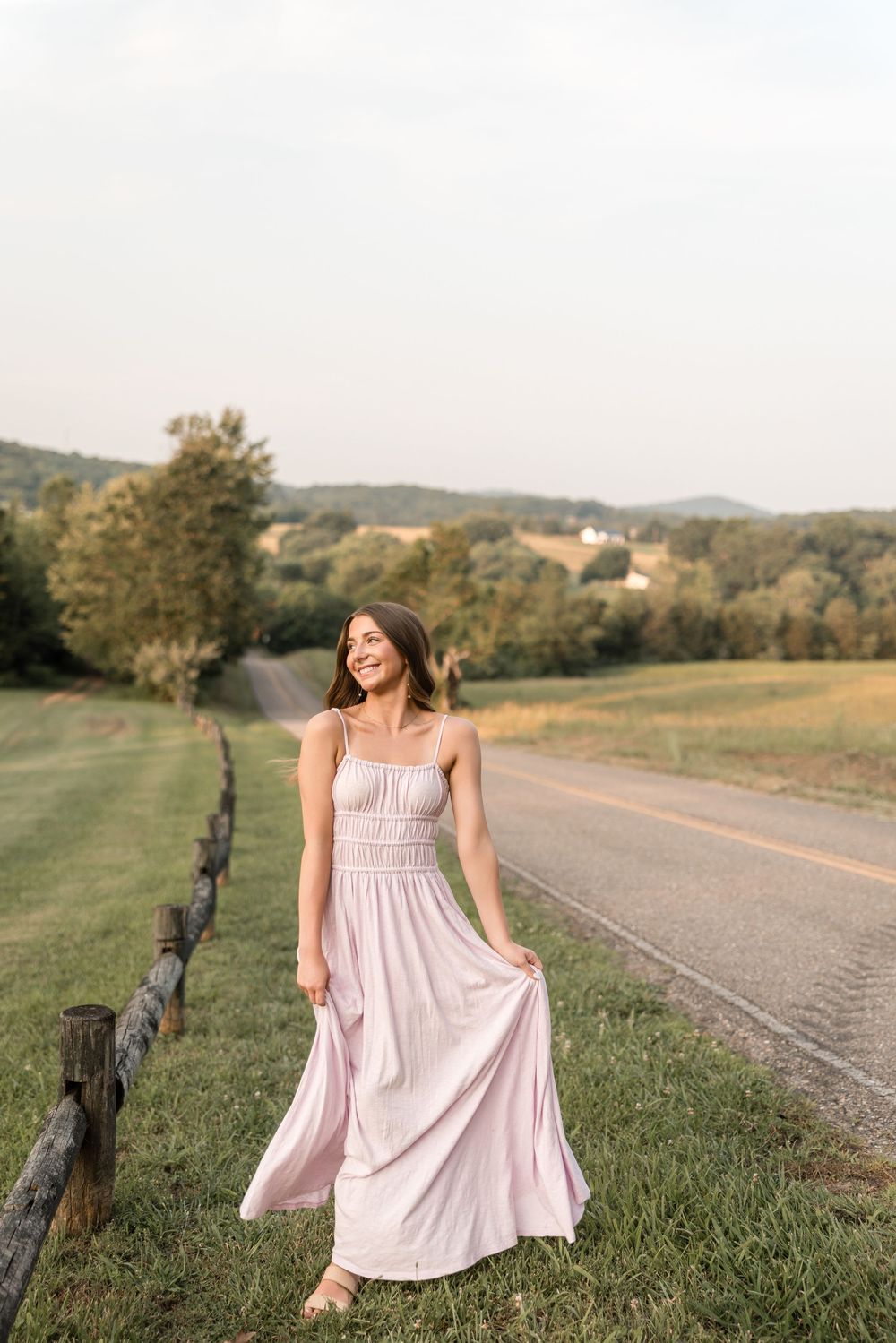 A pink maxi dress flows in the breeze along a rural country road with rolling hills and trees in the background.
