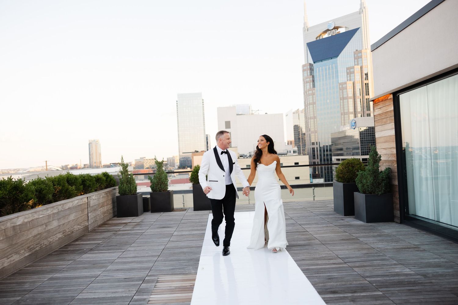 A happy couple in wedding attire runs along a rooftop terrace with a city skyline in the background.