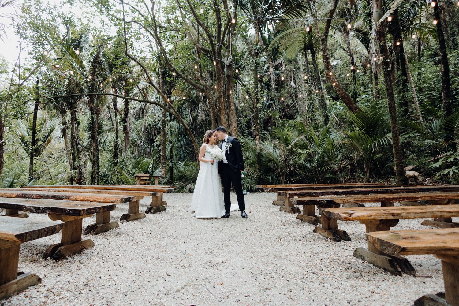 A couple stands between rows of rustic wooden benches captured by Apertured Media Auckland Wedding Photographer.