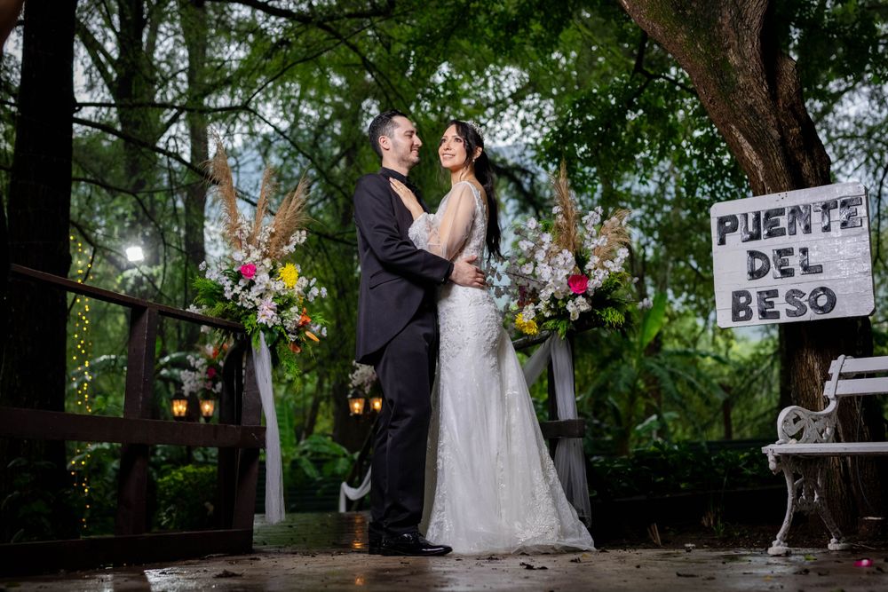 Couple poses under wooden arch with floral decorations in lush garden setting.