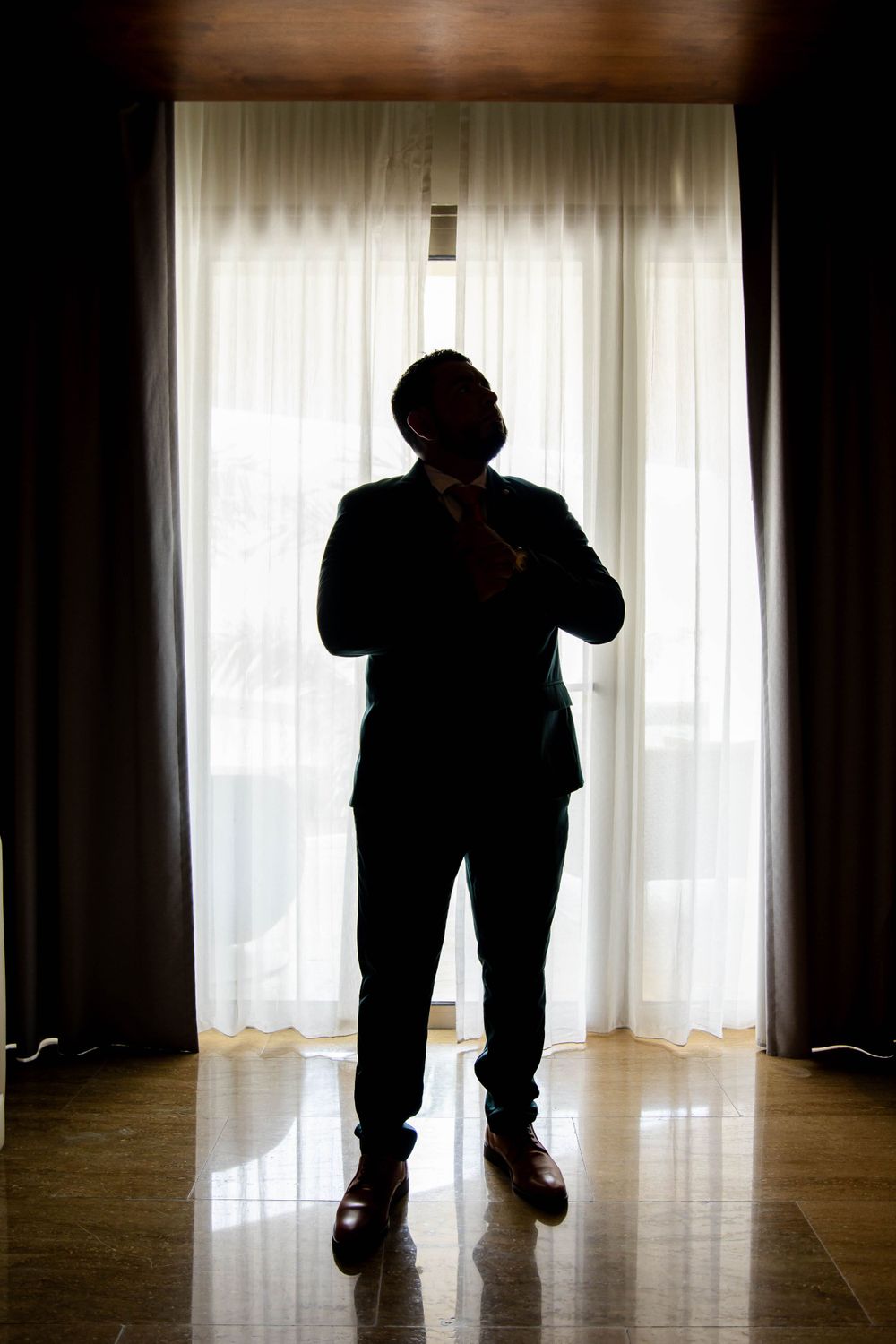 A silhouetted figure adjusts their tie while standing against bright window light.
