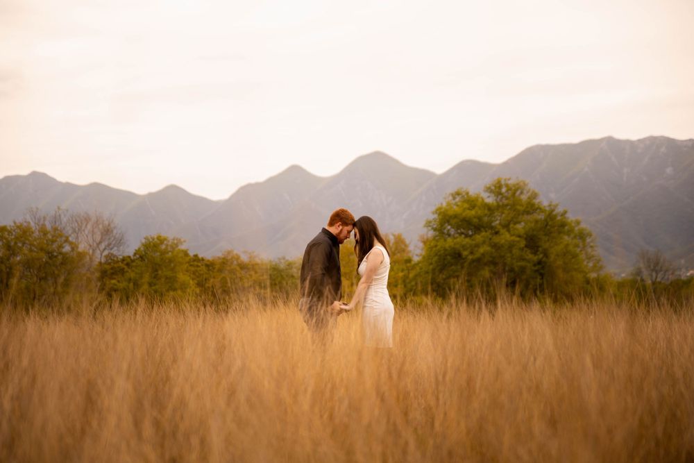 A romantic couple stands together in a golden field with majestic mountains silhouetted in the background at sunset.