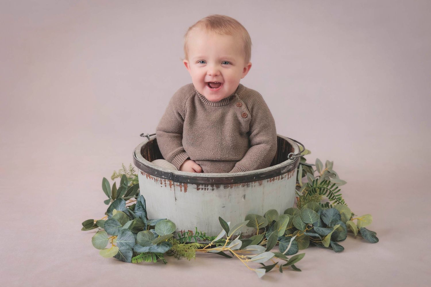 A joyful little one sits in a rustic metal container surrounded by soft greenery. baby milestone tiny moments photography, Gosport Hampshire