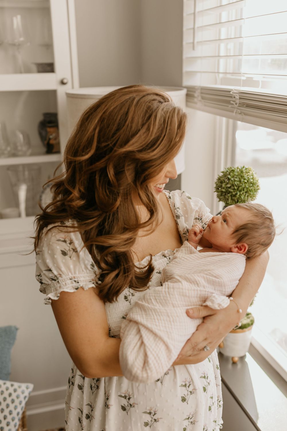 Mother in floral dress holds sleeping newborn by sunny window.