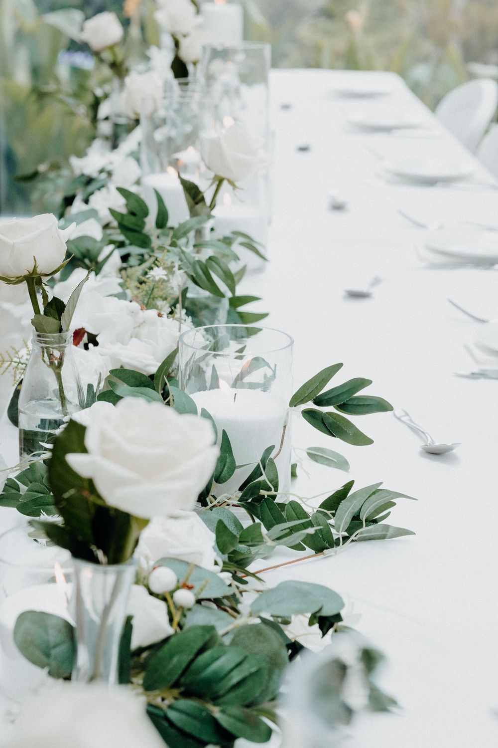 Elegant white roses and eucalyptus leaves decorate a long white wedding table captured by Apertured Media Auckland Wedding Photographer.