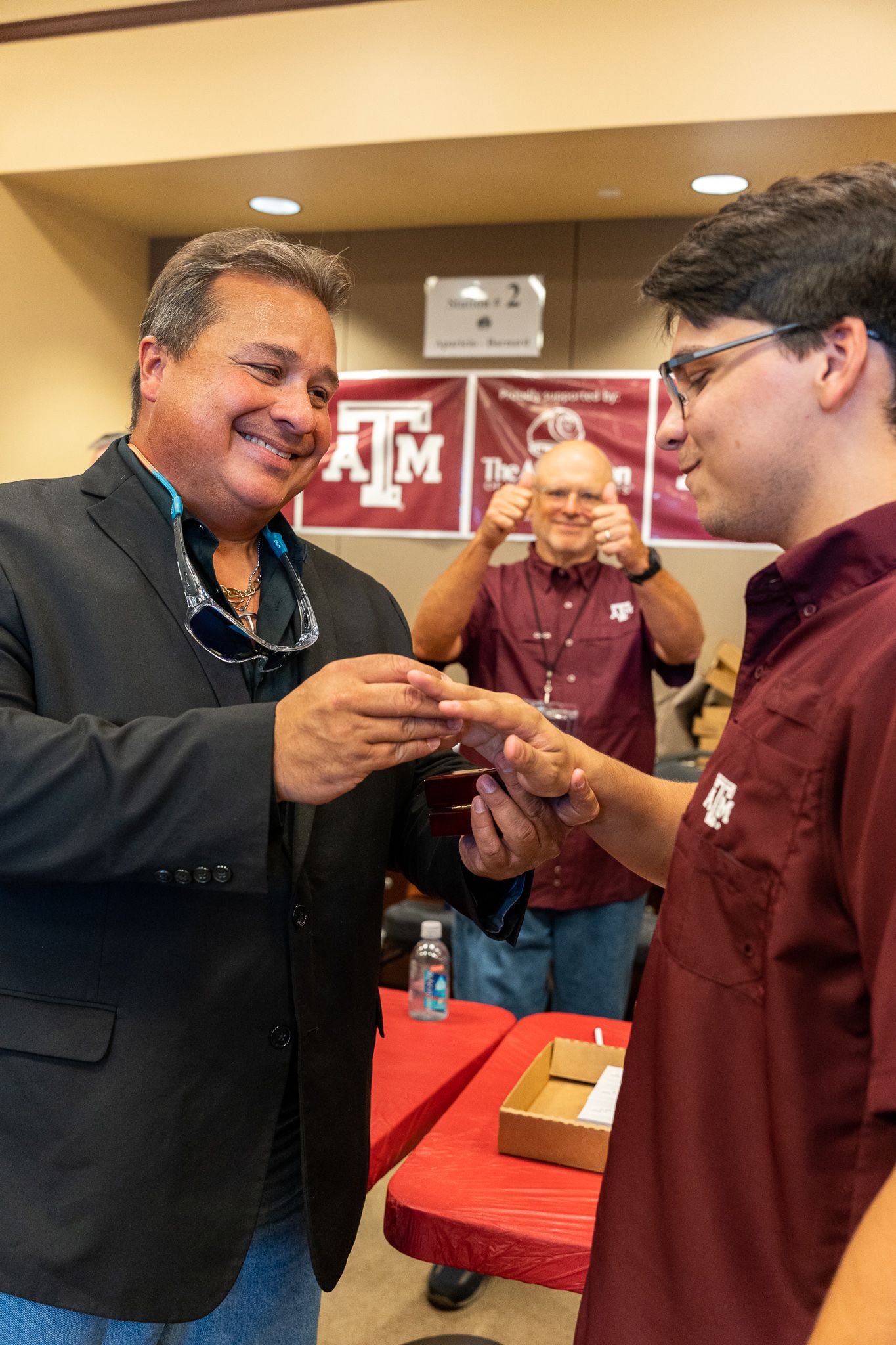 Two people exchange items while a third person in maroon smiles in the background at Texas A&M University.
