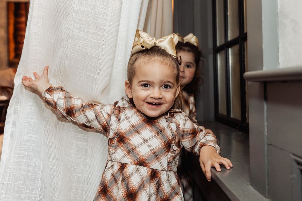 Child in plaid dress playing peek-a-boo behind white curtain.