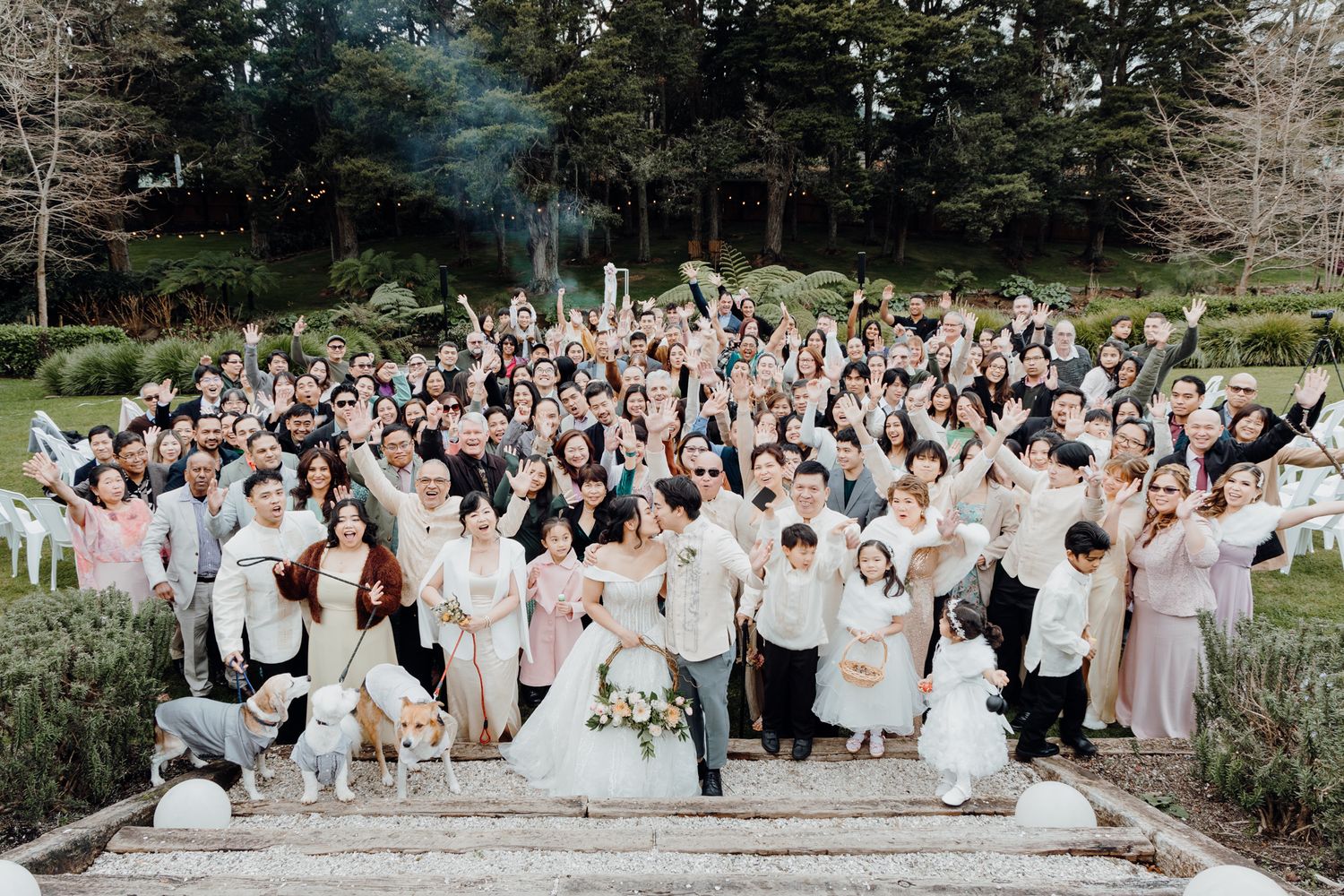 Large wedding group photo with guests dressed in white and pink gathered on outdoor steps.