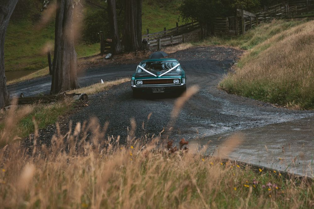 Classic teal car driving on a scenic mountain road surrounded by tall grass.