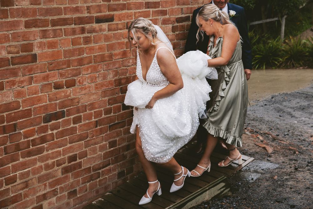 Two women in wedding attire lean against a brick wall while holding their dresses up to display their footwear.