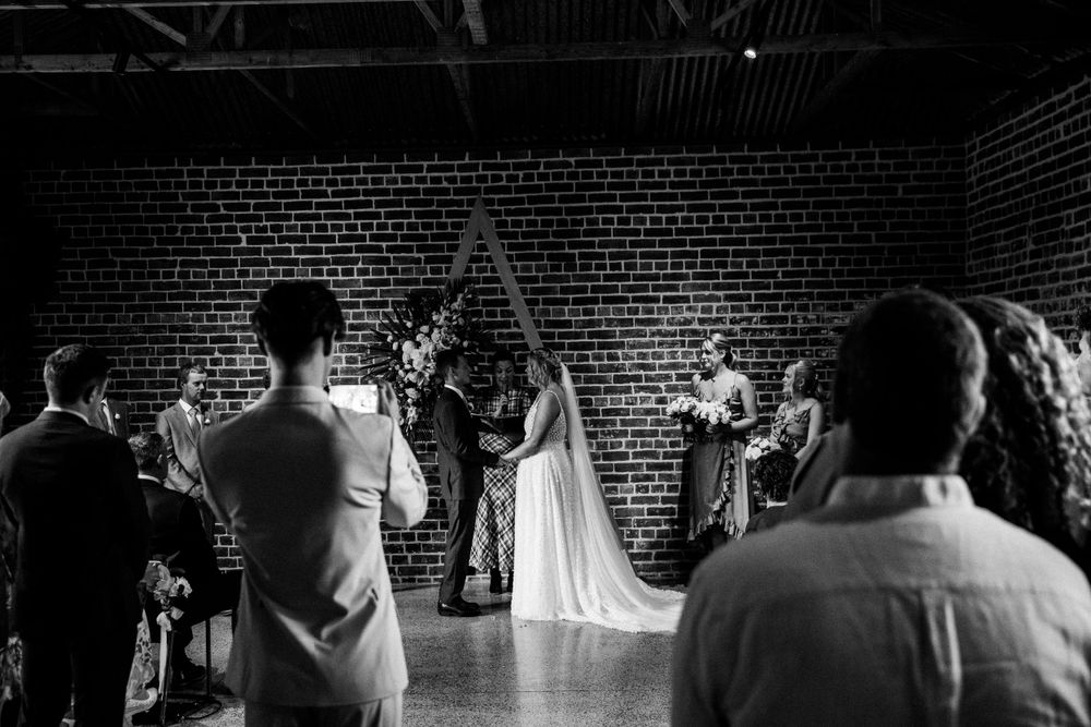 A black and white wedding ceremony photo captured in an industrial brick venue with dramatic lighting.