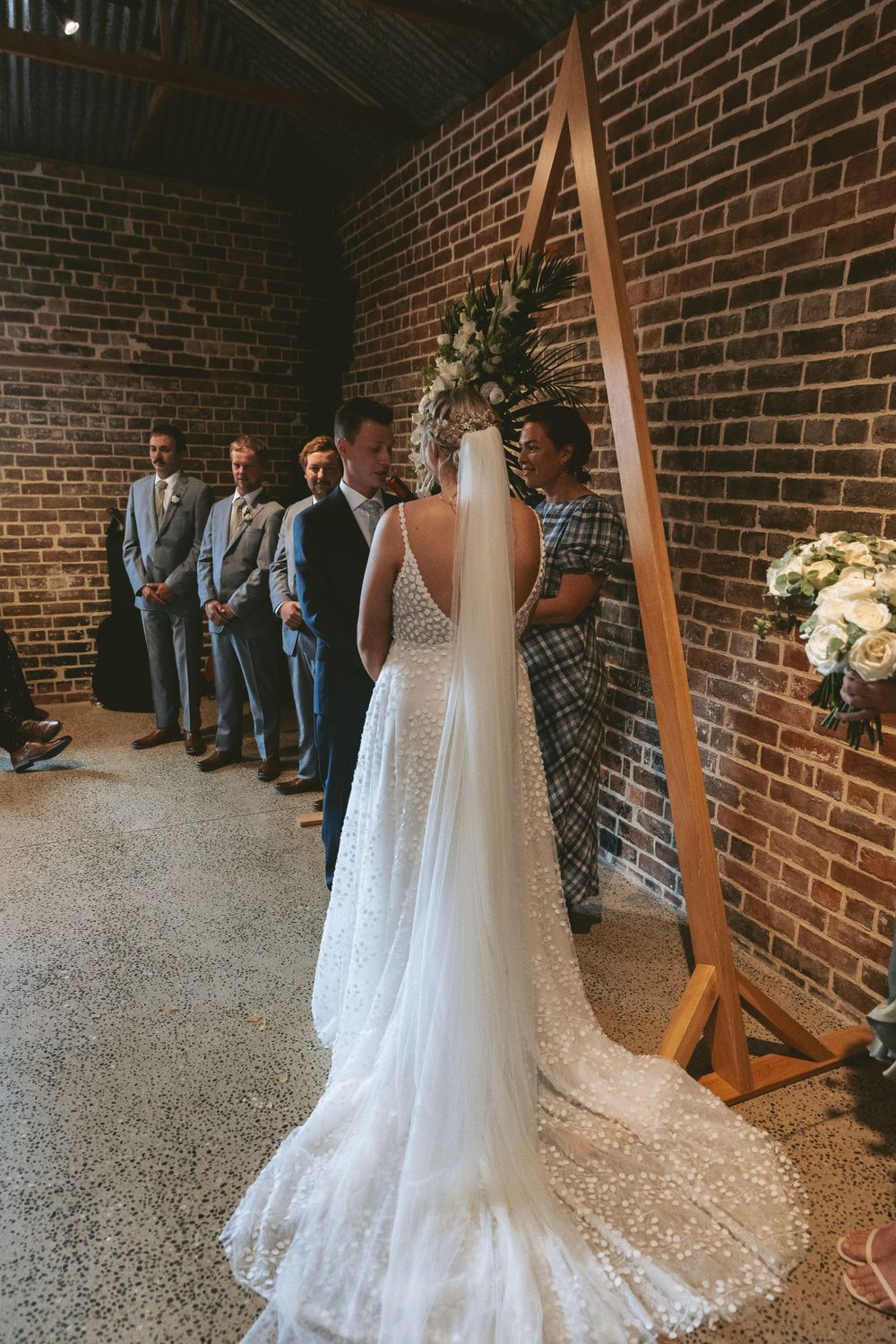 A wedding ceremony in progress with a wooden triangle arch and floral arrangements against brick walls.