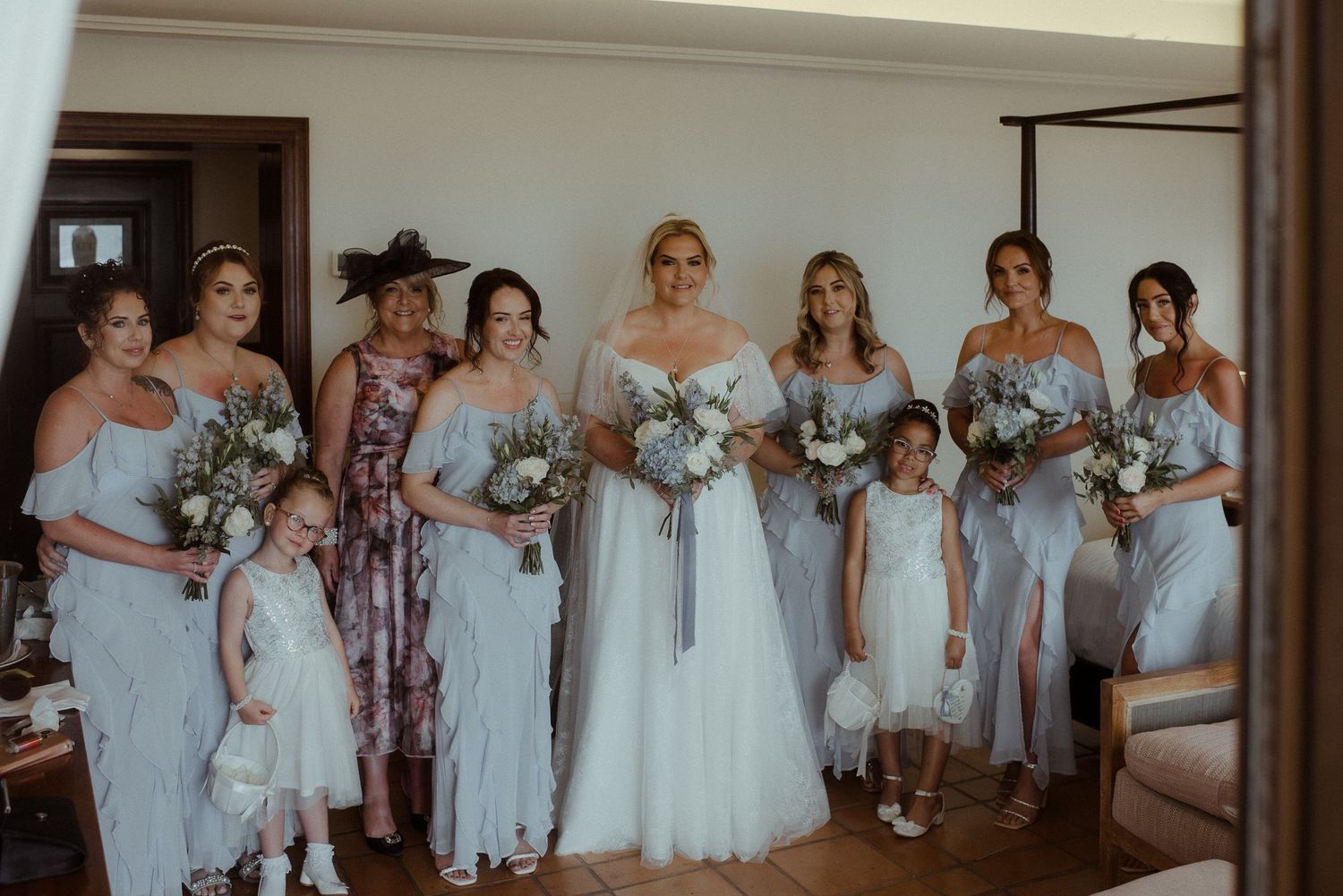 Wedding party poses together in coordinated light colored dresses with white and purple bouquets.