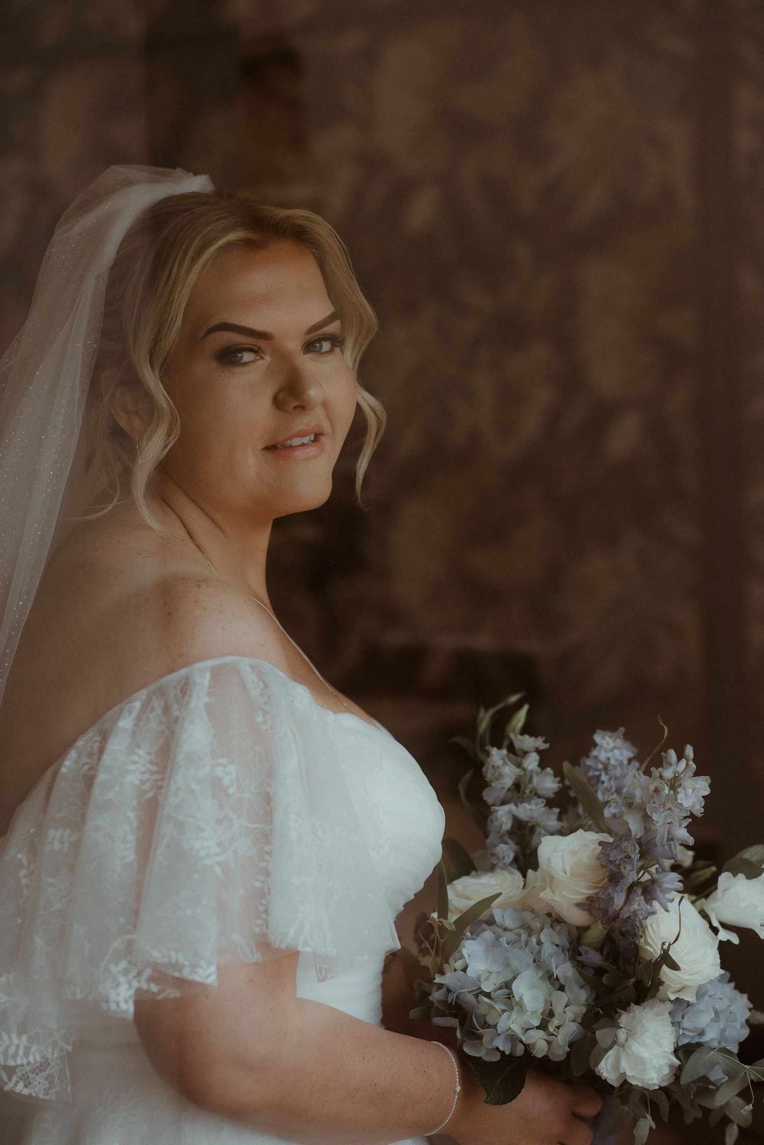 Bride in an off-shoulder wedding dress with delicate lace details holds a white and purple bouquet.