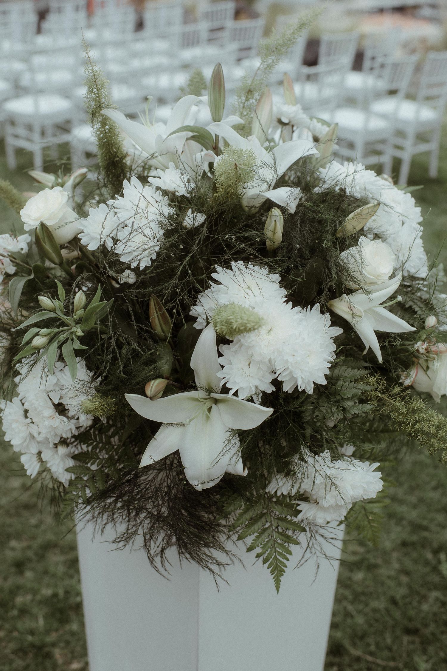 An elegant white floral arrangement with lilies and carnations in a modern white pedestal vase.