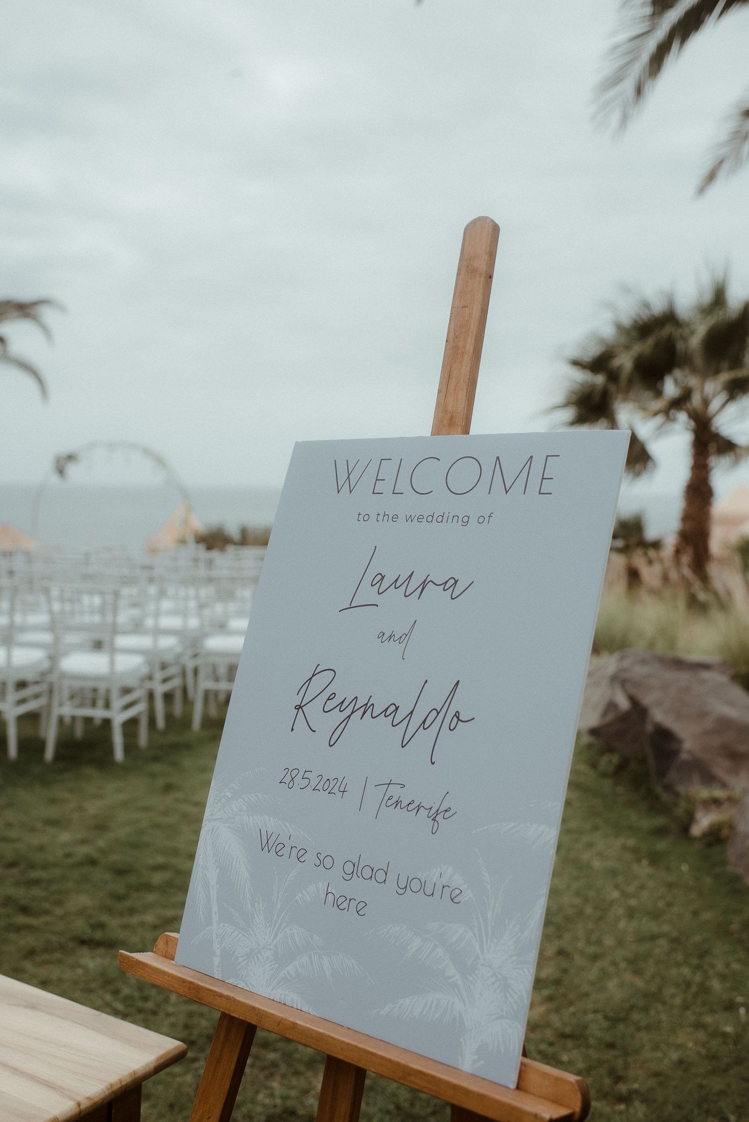 A wedding welcome sign on an easel displaying Laura and Reynaldo's names with white chairs in the background.