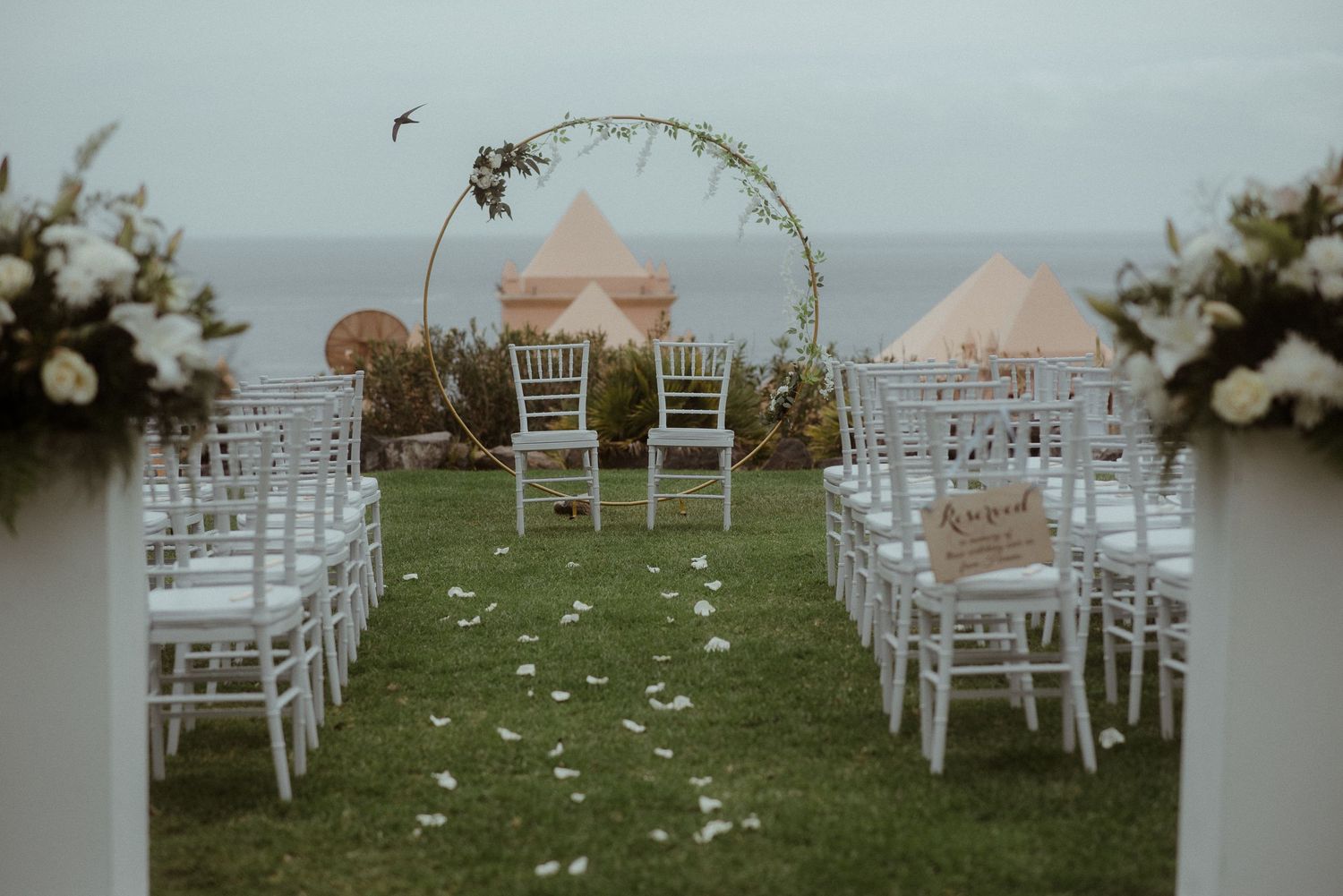 A romantic outdoor wedding ceremony setup with white chairs and a floral arch overlooking the ocean.