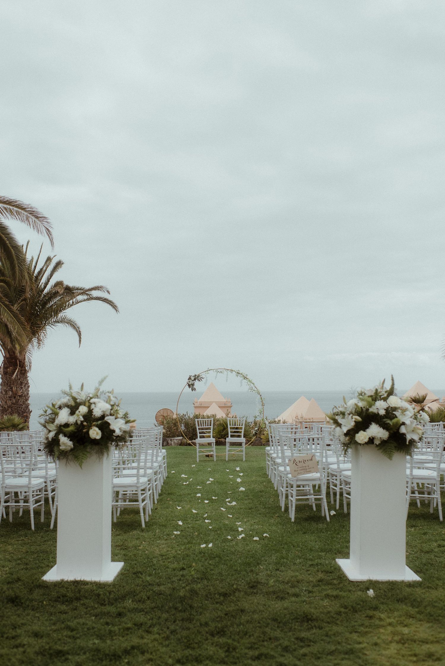 An elegant outdoor wedding venue with white columns, floral arrangements, and palm trees by the ocean.