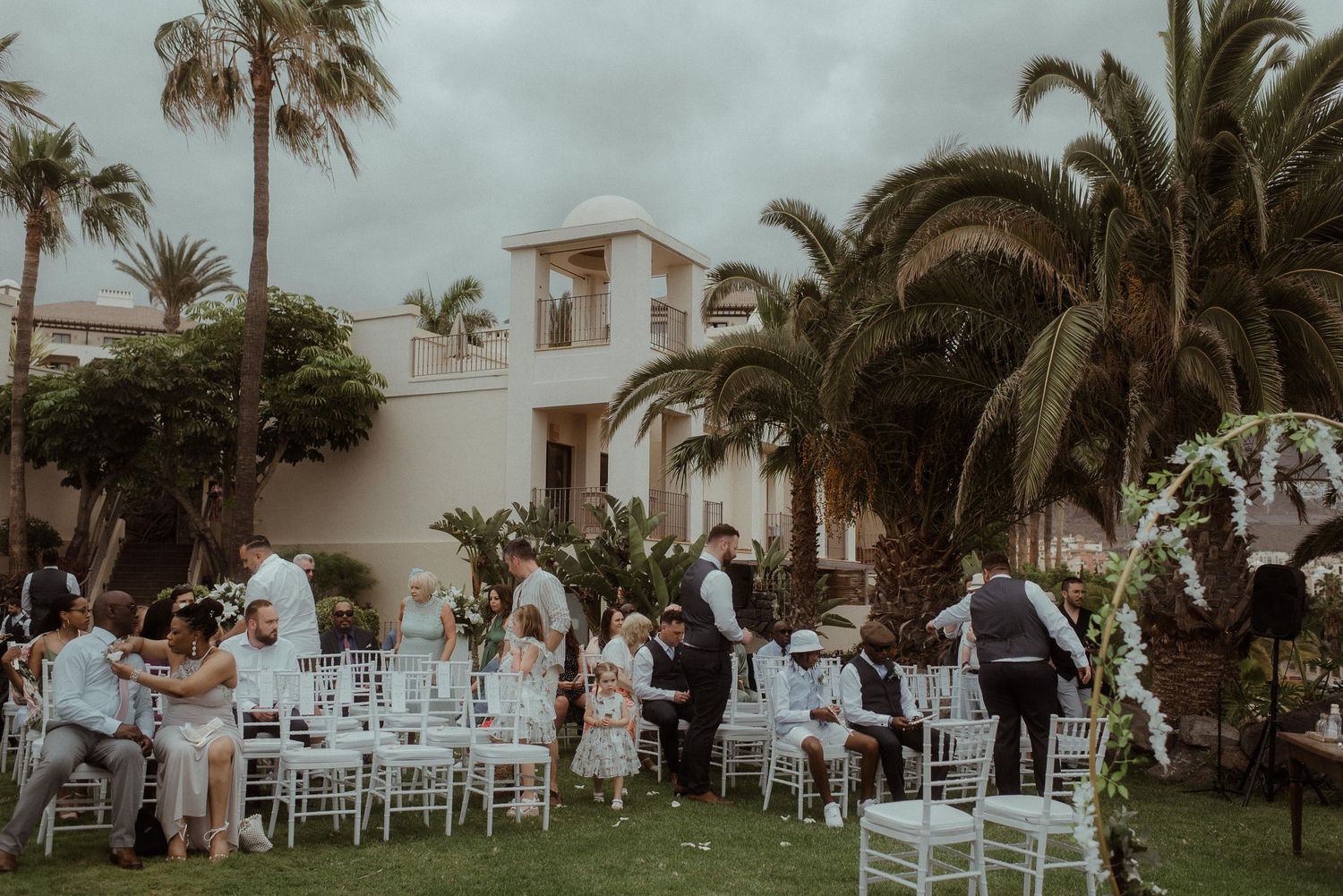 An outdoor wedding reception at dusk with white chairs and palm trees surrounding a Mediterranean-style venue.