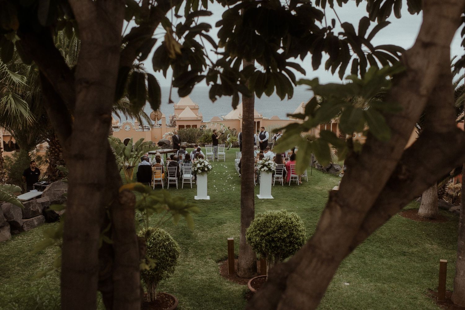 A garden wedding reception viewed through tree branches with guests seated at round tables.
