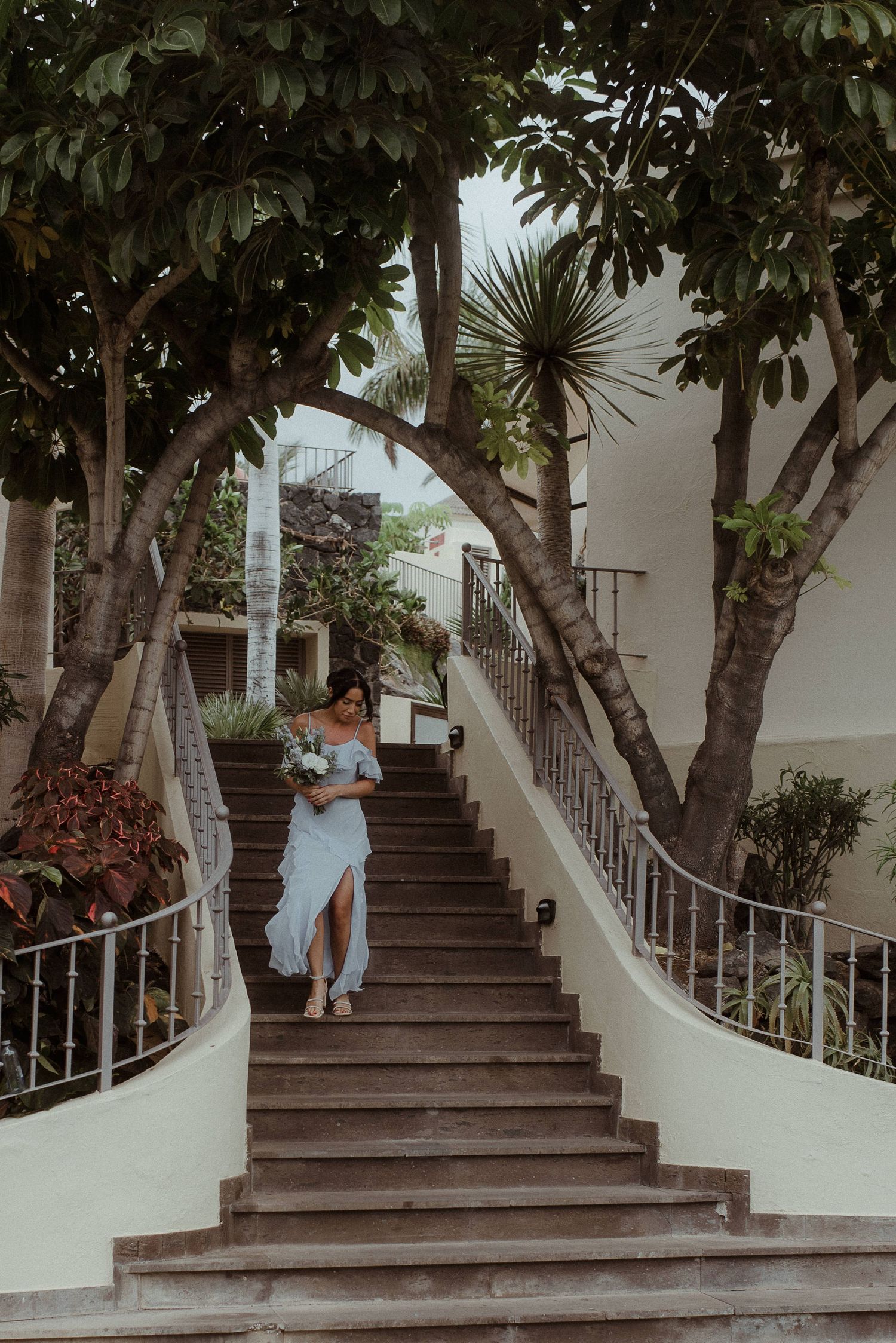 A bridesmaid descending an elegant white curved staircase surrounded by tropical trees.