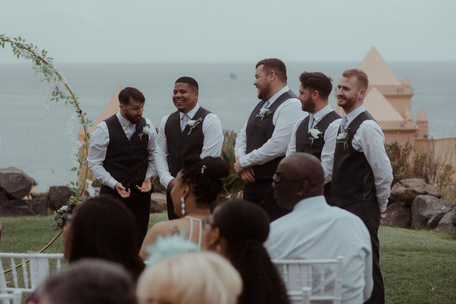 Groomsmen in matching gray vests share a laugh at an outdoor wedding ceremony by the ocean.