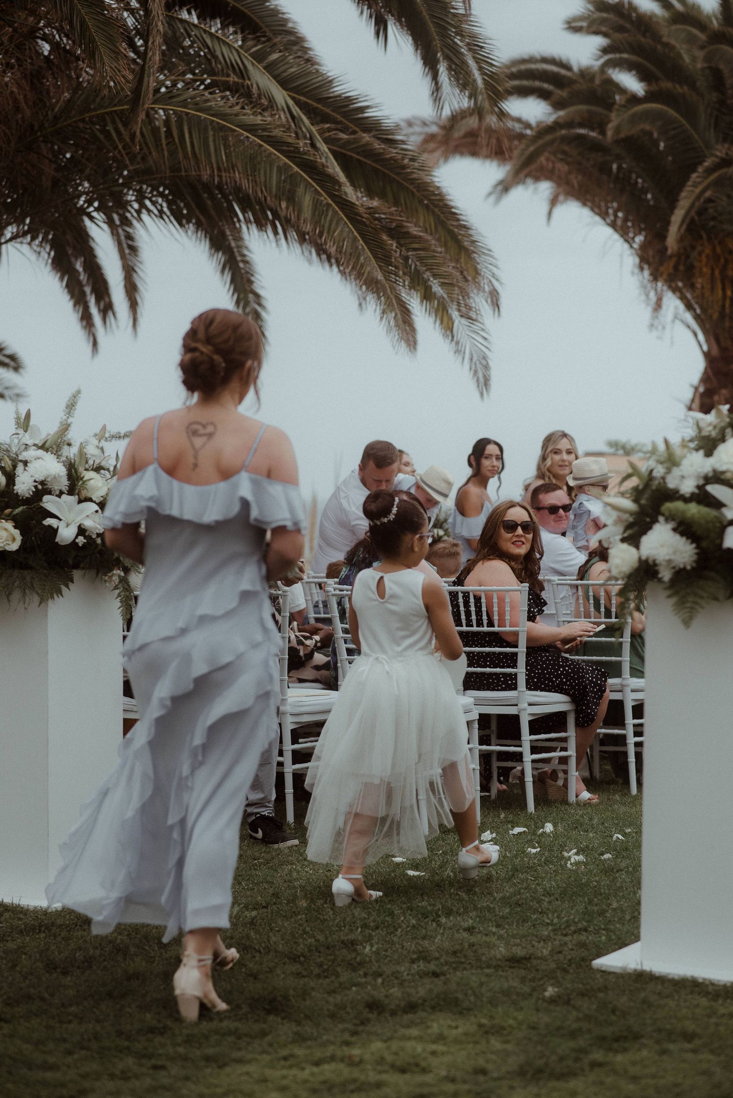 Wedding guests in formal attire seated under palm trees during an outdoor ceremony with white floral decor.