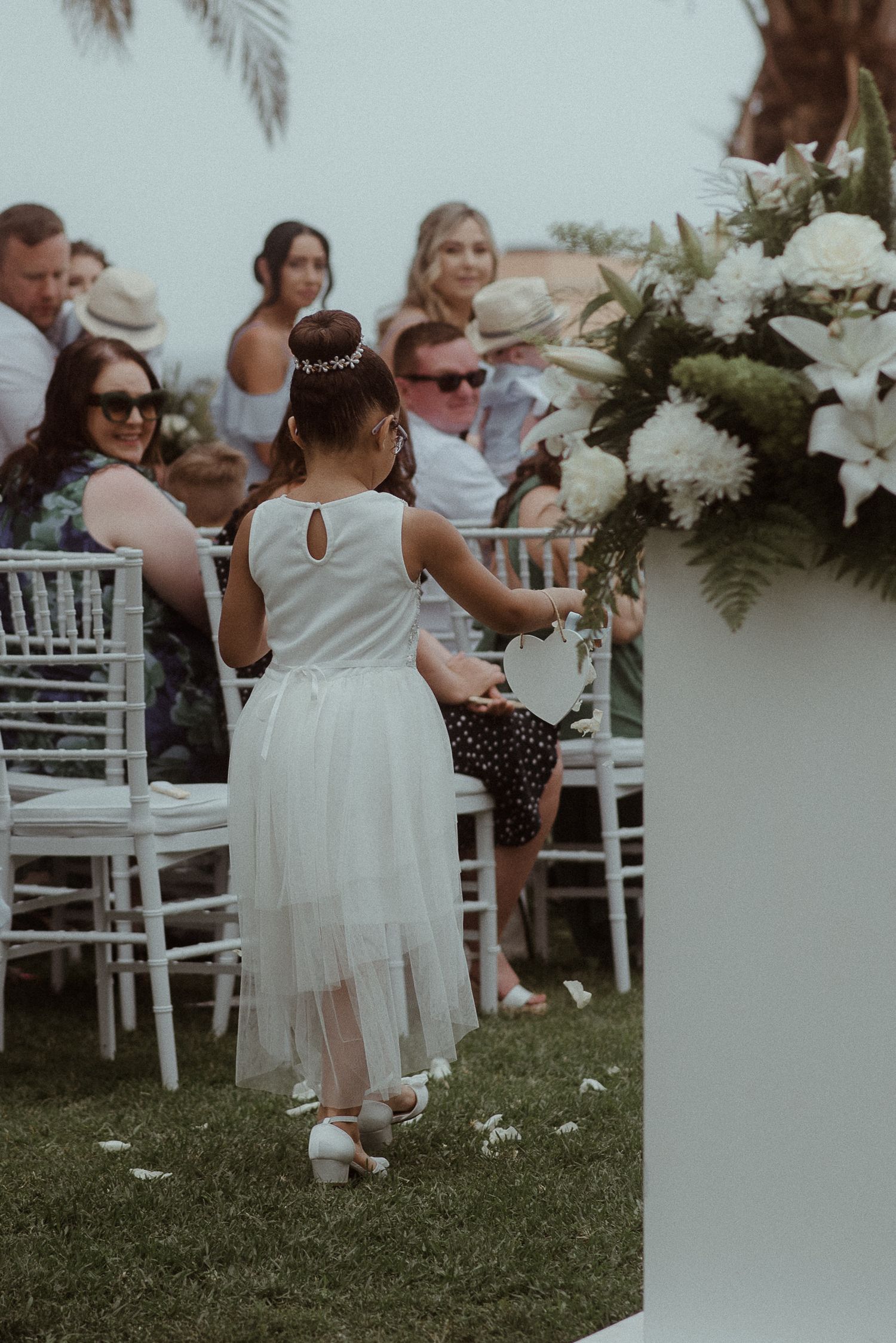 A child in a white dress explores floral arrangements during an outdoor wedding ceremony.