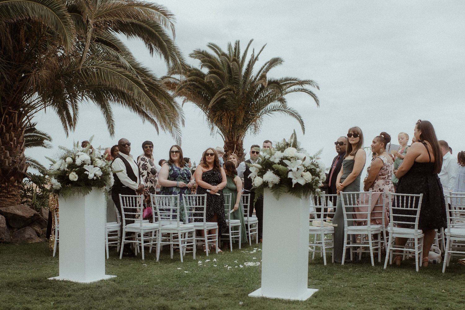 Guests stand between white floral pillars at an outdoor wedding ceremony with palm trees in background.