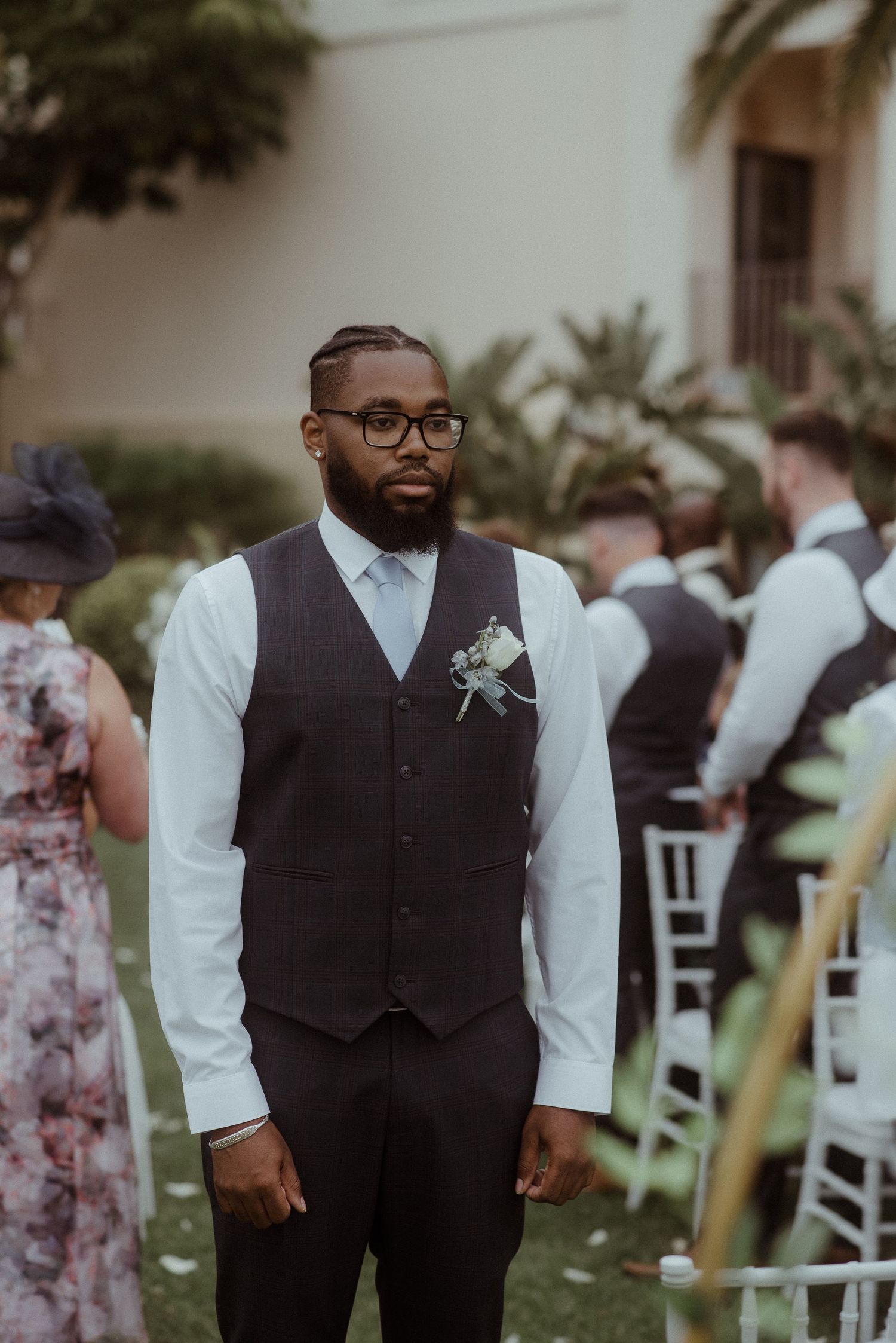 A groomsman in a gray vest and glasses stands at attention during an outdoor ceremony. Tenerife Wedding Photographer at Hotel Vincci